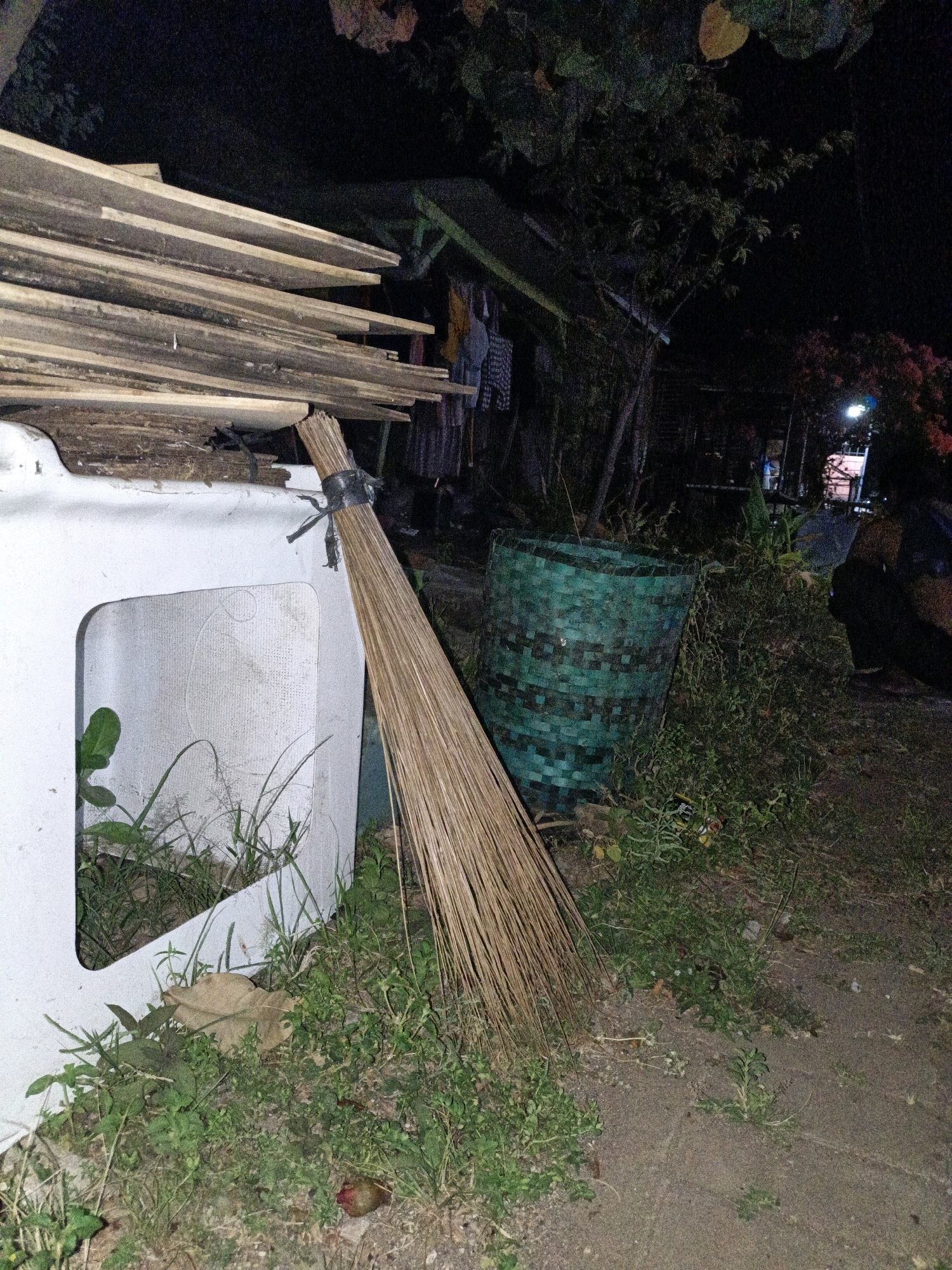 A wide shot taken at night in what appears to be an outdoor area in Cikal, Indonesia, shows a stack of light-colored wooden planks leaning against a white, upright object with an open rectangular section. A broom made of dried grass is tied with black tape and leans against the white object, with its bristles touching the ground. To the right of the broom and white object is a tall, cylindrical basket woven from dark green material. Beyond the basket, there is dense vegetation, including bushes and trees. In the background, a small building with a dark roof and some clothing hanging on a line is visible. A bright light illuminates part of the background, where a person in dark clothing can be seen standing. The ground is a mix of dirt and sparse grass, with some fallen leaves and debris.