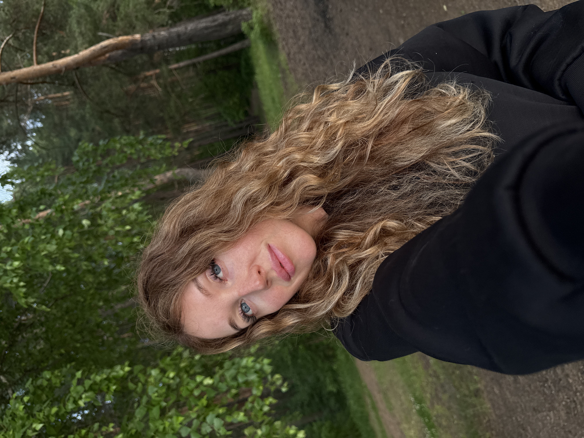 A young woman with long, wavy blonde hair is taking a selfie outdoors in a wooded area, likely in or near the city of Chaikovsky, Russia. She is wearing a black hooded sweatshirt and is looking towards the camera with blue eyes and pink lips. The background consists of green foliage and trees, suggesting a natural setting like a park or forest.