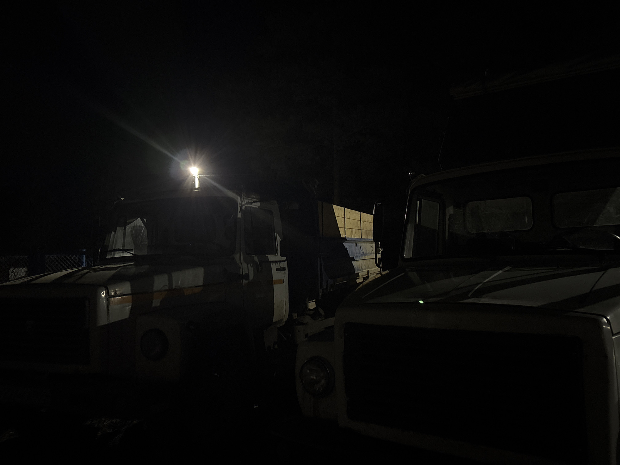 The image depicts two vehicles, possibly trucks, parked closely together in a dimly lit environment, likely at night. The location is unknown. The trucks appear to be older models, with boxy designs. The scene is dark, obscuring many details of the vehicles and their surroundings. Only the front portions of the trucks are clearly visible. A light source, possibly from one of the trucks, illuminates the front of the vehicle on the left.