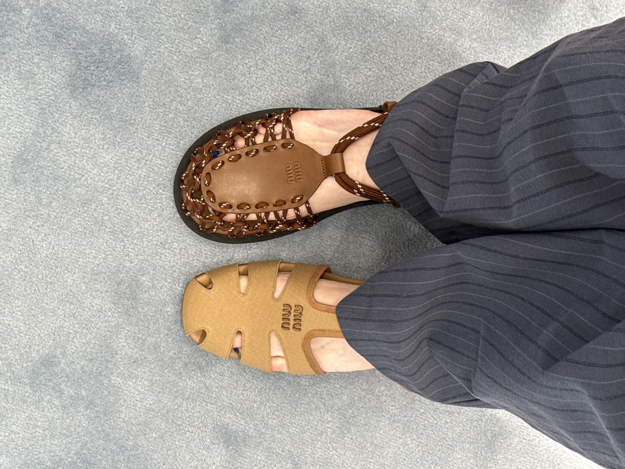 The image shows a person wearing mismatched Miu Miu sandals while standing on a light blue carpet or rug. The sandal on the left foot is brown and beige with a woven strap design, while the sandal on the right foot is light brown with cut-out details. The person is wearing dark blue pinstripe pants. There is no indication of the image being taken in Costa Mesa, United States of America.