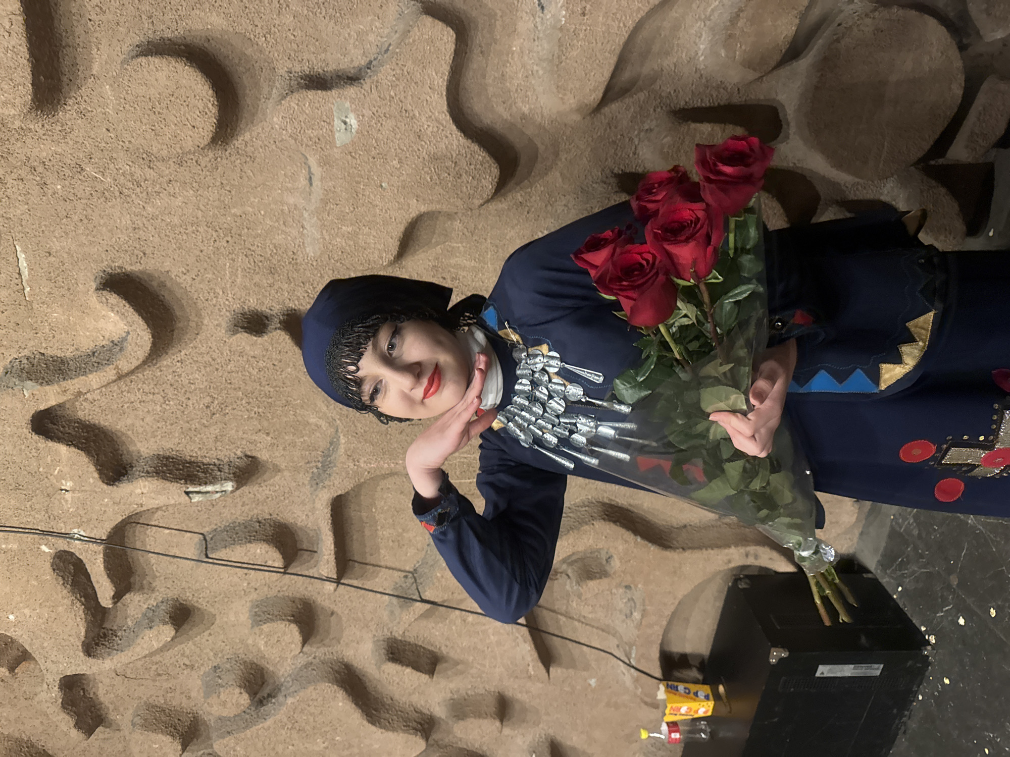 A woman in traditional Georgian dress stands in front of a textured wall, possibly a replica of ancient architecture, in what appears to be an indoor setting, potentially in Tbilisi, Georgia, given the traditional Georgian attire. She wears a dark blue outfit with silver ornamental jewelry and a matching head covering. She is holding a bouquet of red roses wrapped in clear plastic. Her right hand is resting under her chin, and she is smiling. To the right of the woman, on the floor, is a black box with several food packets on top of it. There is a wire going across the background behind her. The scene is well-lit.