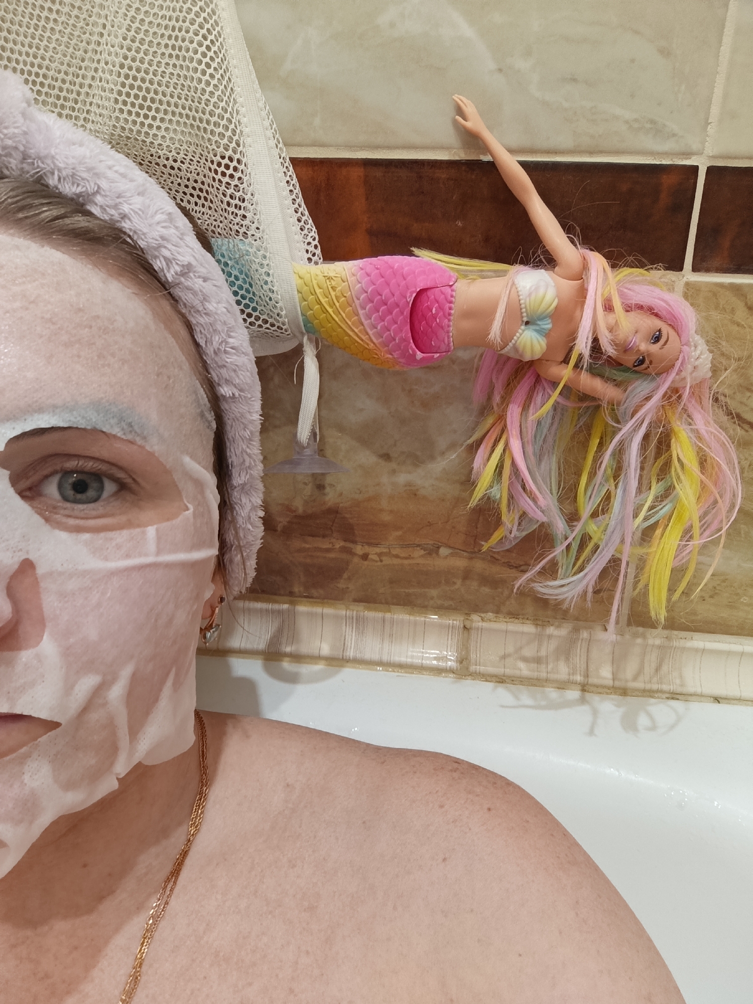The image captures a close-up, vertical view of a bathroom scene, likely in Chernogolovka, Russia, from the perspective of someone in a bathtub.
The primary subject is a person, visible from the chest up on the left and bottom of the frame. Their face is mostly covered by a white sheet mask, with their left eye peering out towards the viewer. They wear a soft, light-colored, fuzzy headband around their head. A thin, golden chain necklace is draped around their neck and visible on their bare chest and shoulder, which has freckles. Their visible eye is light-colored, possibly blue or green.
To the right, a colorful mermaid doll is suspended horizontally against the tiled wall. The doll features long, flowing hair in shades of pink, yellow, blue, and purple, and a shimmering, multi-colored (pink, yellow) mermaid tail. It wears a seashell-shaped bra in white, yellow, and blue. The doll is attached to a white mesh bag, which appears to be a bath toy organizer or shower caddy, hanging from the wall via a clear suction cup.
The background consists of light beige and darker brown horizontally striped bathroom tiles. A section of a white bathtub rim is visible along the bottom edge.
The activity taking place is a personal relaxation or self-care moment, with the person applying a face mask, likely while bathing. The mermaid doll adds a playful or decorative element to the bathroom setting. There is no visible text in the image.