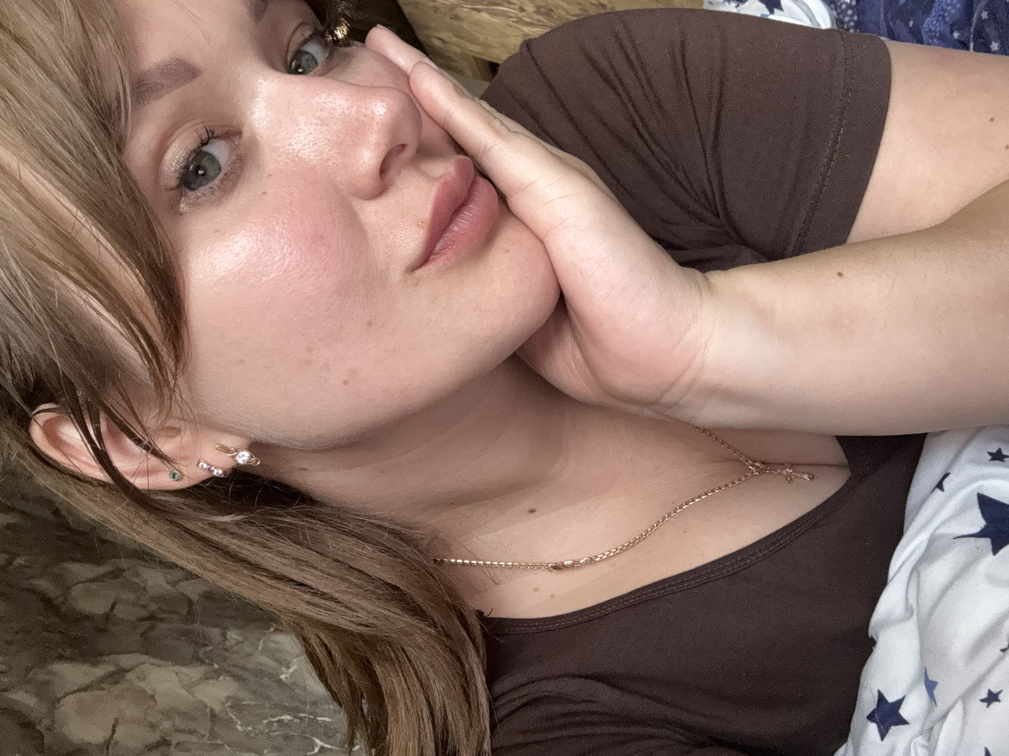 The media file contains a close-up, head-and-shoulders shot of a young woman with light brown hair and fair skin, lying down and resting her head on her hand. She is wearing a brown t-shirt and a delicate gold necklace with a small cross pendant. Her makeup is subtle, with emphasis on her eyes and lips. She has multiple earrings in her left ear, including a stud with a green stone and a stud with a white stone and pink accents. The background is out of focus, with hints of a textured surface to her left and patterned bedding to her right, featuring blue and white stars. The lighting suggests an indoor setting, likely during the day. Her expression appears relaxed and contemplative. No visible text is present in the image. The provided location context is Chaikovsky, Russia.