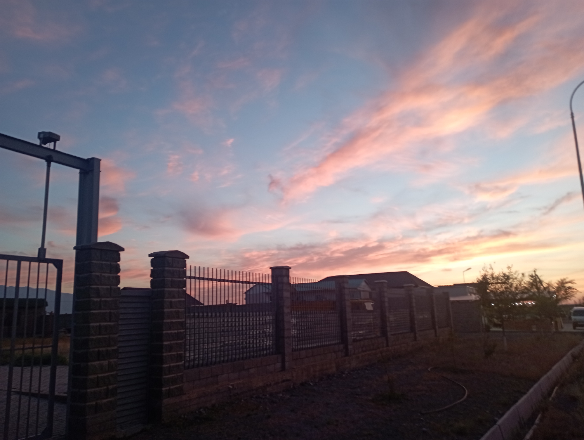 A wide-angle shot captures a sunset over a residential area. The sky is a soft blend of blue and pink, with wispy clouds stretching across. In the foreground, a dark metal gate stands on the left, leading to a brick fence with metal bars. Beyond the fence, several houses with dark roofs are visible. A street lamp curves over the scene on the right, and a white vehicle is parked further down the street. The ground is covered in dark gravel.