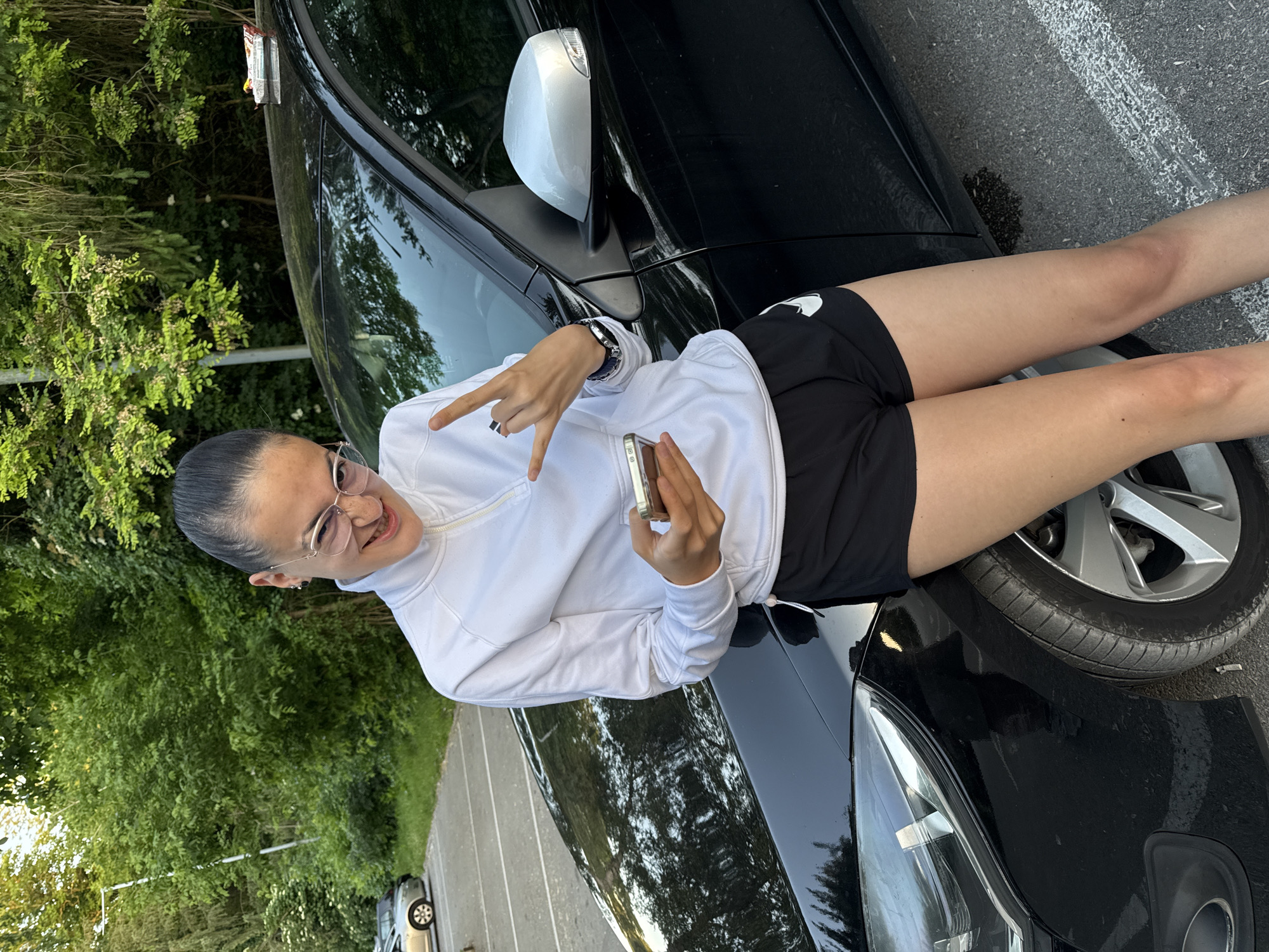 A woman is sitting on the hood of a black car. She is wearing a white zip-up sweater and black shorts with a visible white logo on the left leg. She is holding a phone in her right hand and making a hand gesture with her left hand. She has her hair pulled back and is wearing glasses. The car is parked on the side of a road with trees visible in the background. The location is in the city of Pleven, Bulgaria.