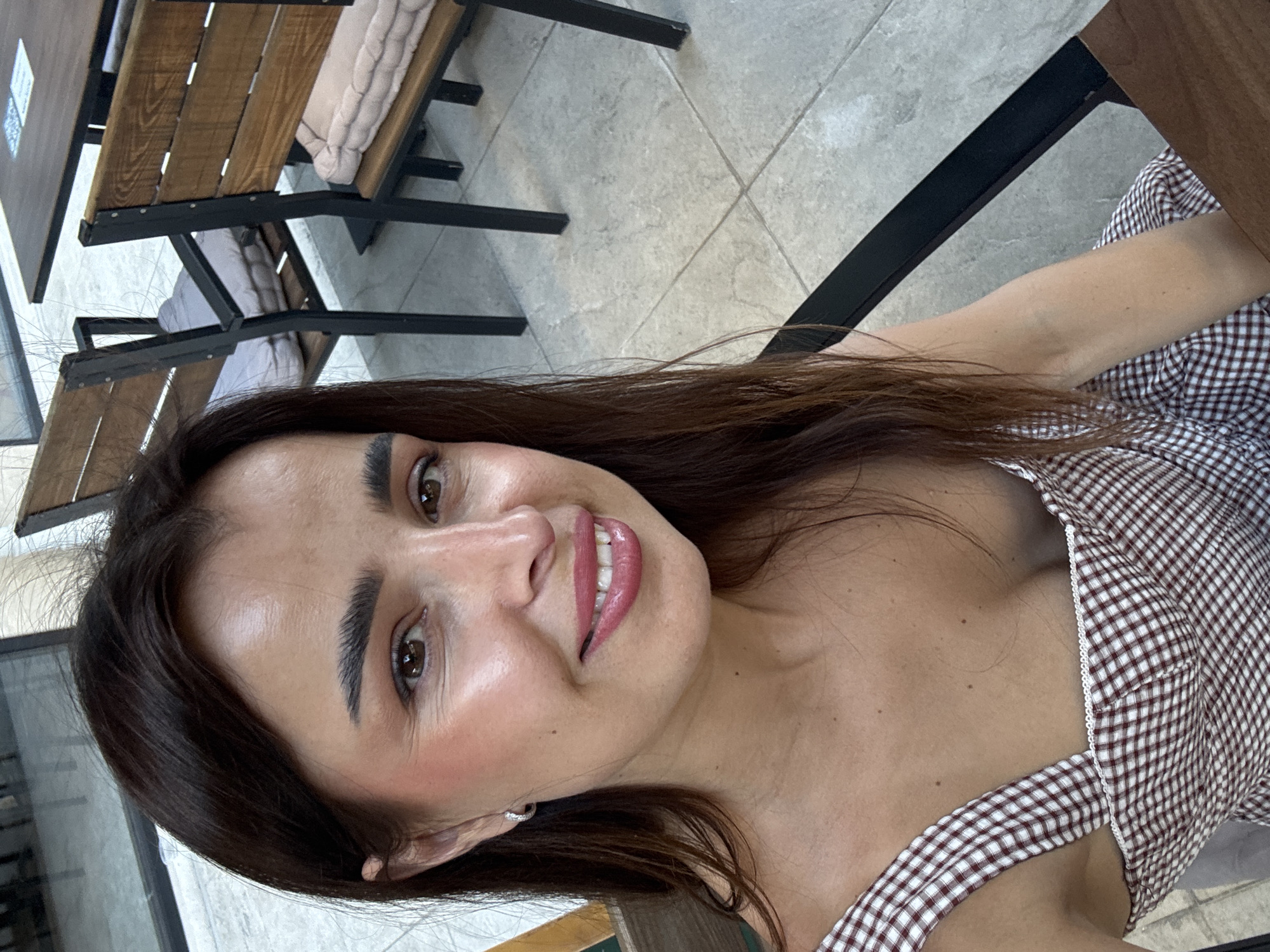 A woman with long, dark brown hair is taking a selfie. She is wearing a brown and white checkered dress. Her makeup includes winged eyeliner and pink lipstick. Her head is tilted to the left, and she is smiling. In the background, there are two wooden chairs with light-colored cushions and a dark wood table, suggesting an outdoor seating area of a cafe or restaurant. The flooring appears to be tiled. The scene is likely in Podgorica, Montenegro, based on the context provided.
