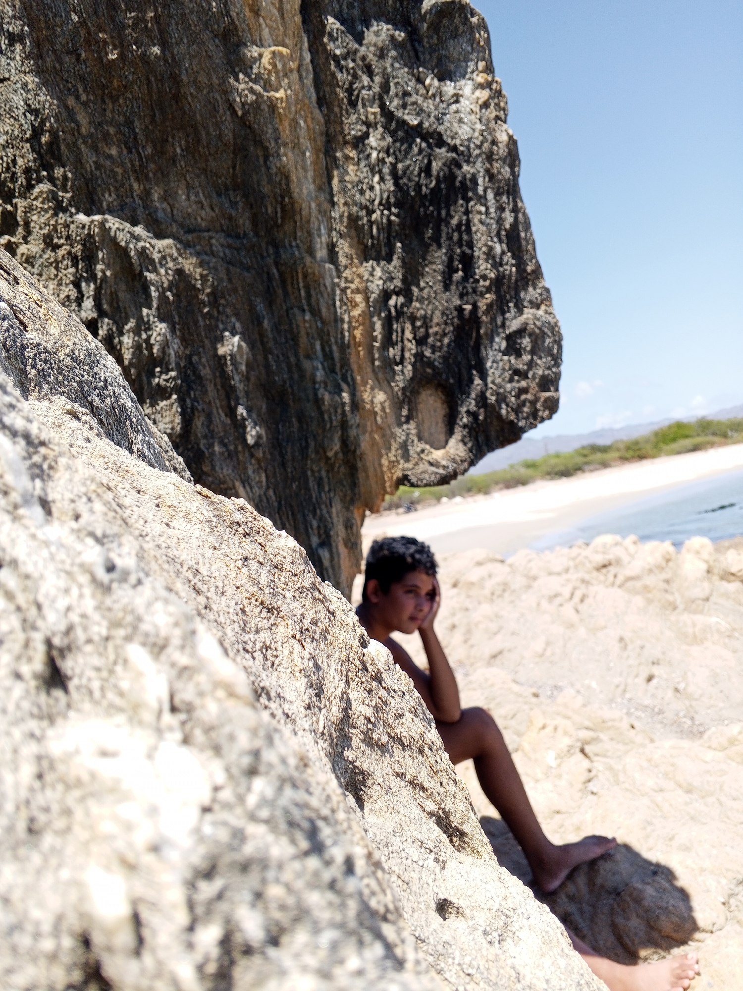 A medium shot captures a young person of color seated on a beach in Juan Griego, Venezuela, leaning against a light-colored rock formation. The individual is partially obscured by the rock, with one hand supporting their head in a pensive posture. A larger, darker rock overhangs the area, creating a shaded space. In the background, a beach with white sand meets a body of water, transitioning to a shoreline dotted with vegetation. The sky is clear and blue.
