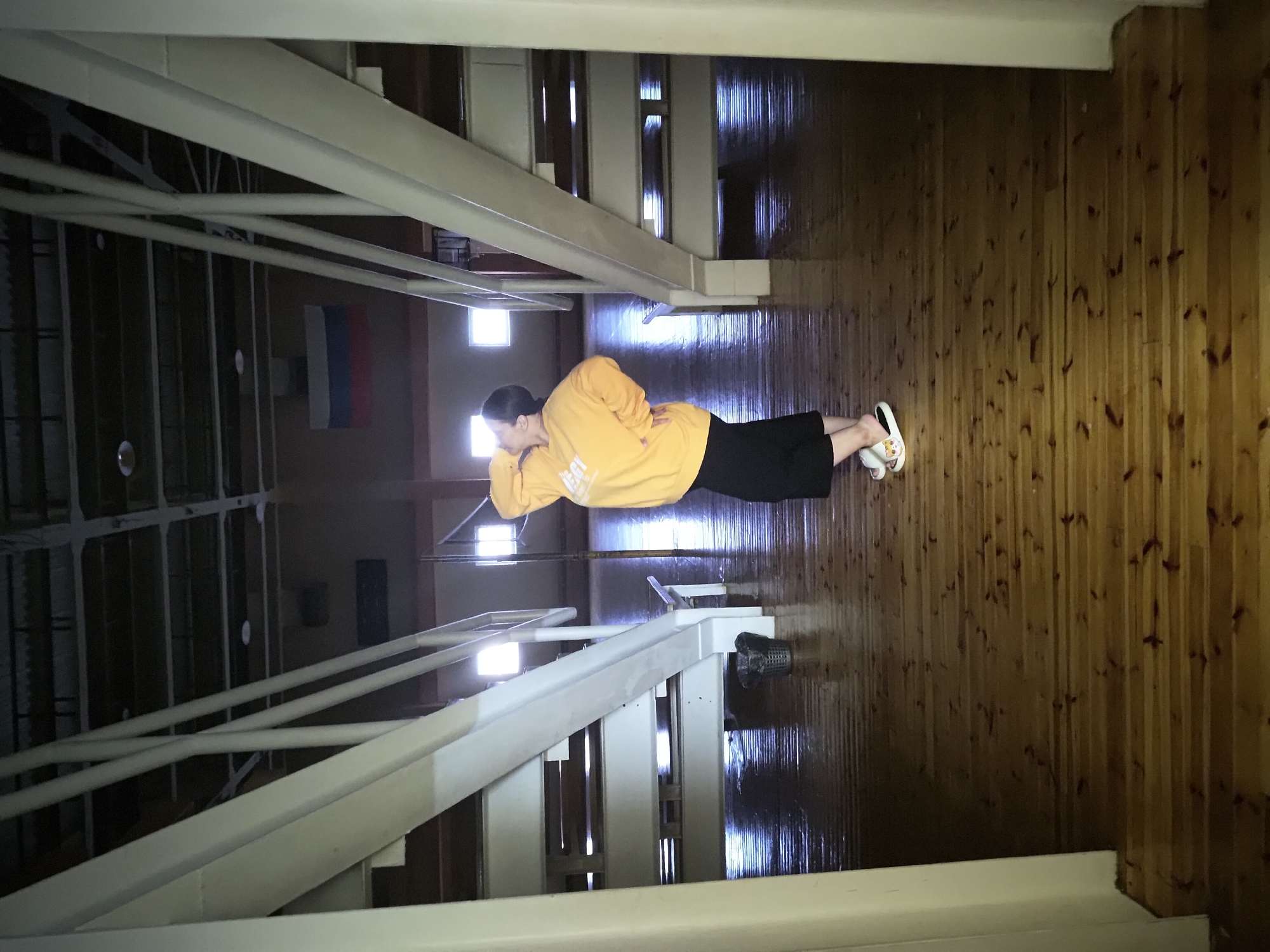 A person wearing an orange sweatshirt and black pants is lying on a glossy wooden floor in what appears to be the gymnasium of a sports facility. The person is lying on their side with their head resting on their hand. White metal staircases are visible on either side of the person. A Russian flag is visible in the background. The location is an indoor space, potentially a gymnasium or similar large hall. The city and specific location are unknown.