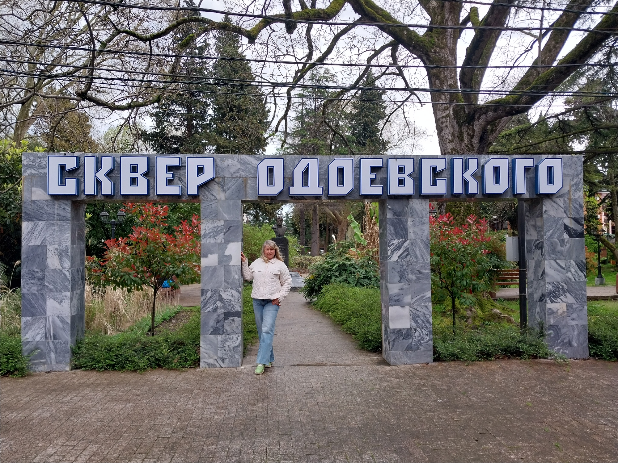 A full, eye-level outdoor shot shows a woman standing in the center of a walkway that leads through a park entrance marked by large, gray stone pillars and the inscription "СКВЕР ОДОЕВСКОГО" in large white Cyrillic letters with blue trim. The park is located in Sochi, Russia.
The woman is blonde and wearing a light cream quilted jacket, blue jeans, and green shoes. She has one hand in her pocket and a slight smile.
The park entrance is made of gray stone blocks arranged in a pillar-and-lintel style. The inscription is on the lintel.
The walkway is paved with patterned gray stones. The park on either side of the walkway is lush with green vegetation, trees, and flowering bushes. A large, old tree with gnarled branches is visible in the background. Power lines crisscross above the park entrance.