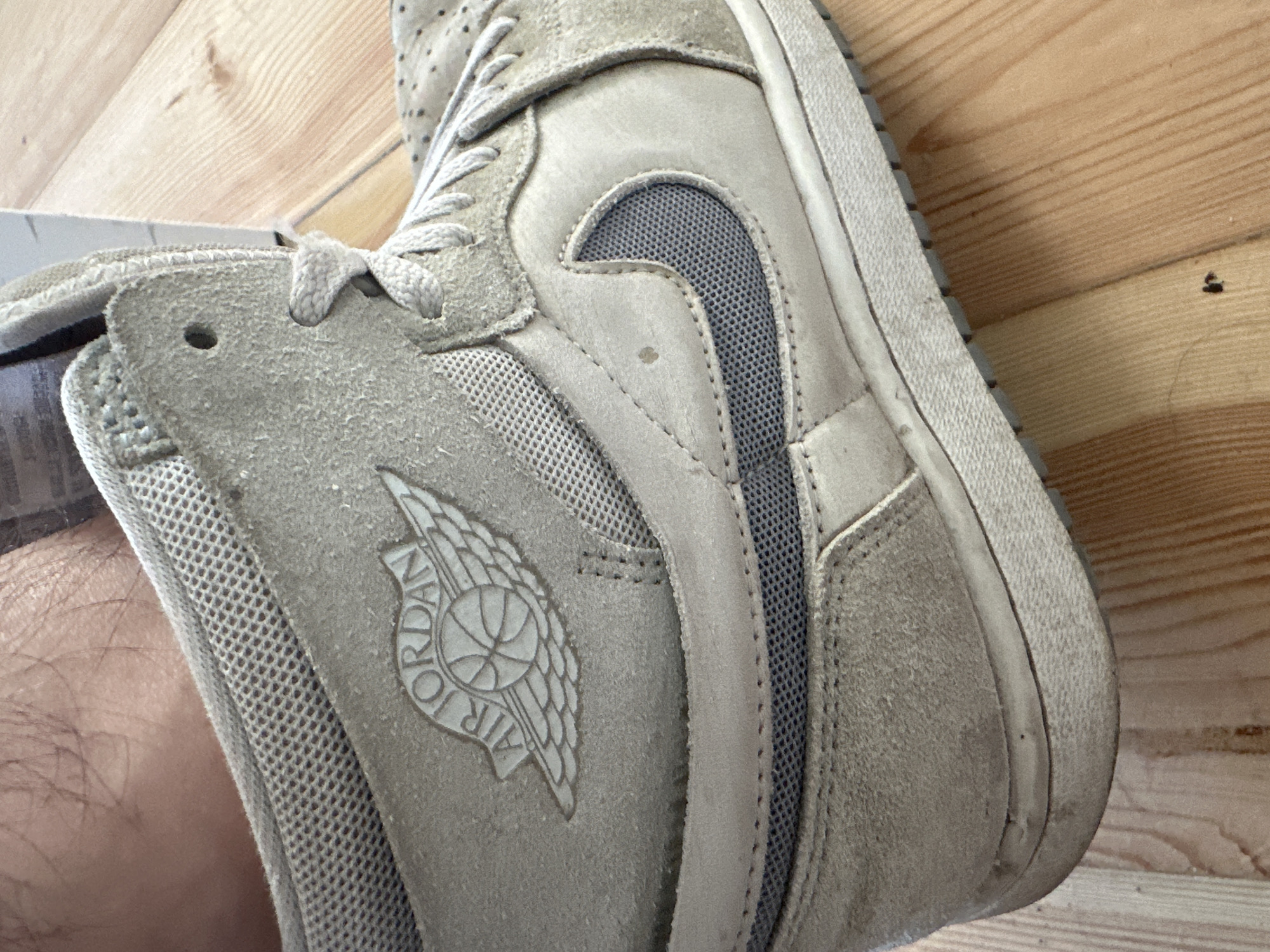 A close-up, medium-shot photograph captures the side of a worn, light beige Air Jordan 1 high-top sneaker resting on a light-brown wooden floor. The sneaker features the iconic Air Jordan Wings logo embossed on its side, a grey Nike swoosh with a mesh backing, and a textured sole. The shoe exhibits signs of use, with dirt and scuff marks visible, particularly on the sole and upper. The background is out of focus, but the wooden floorboards are clearly discernible. No specific location in Łódź, Poland, can be identified from the image.