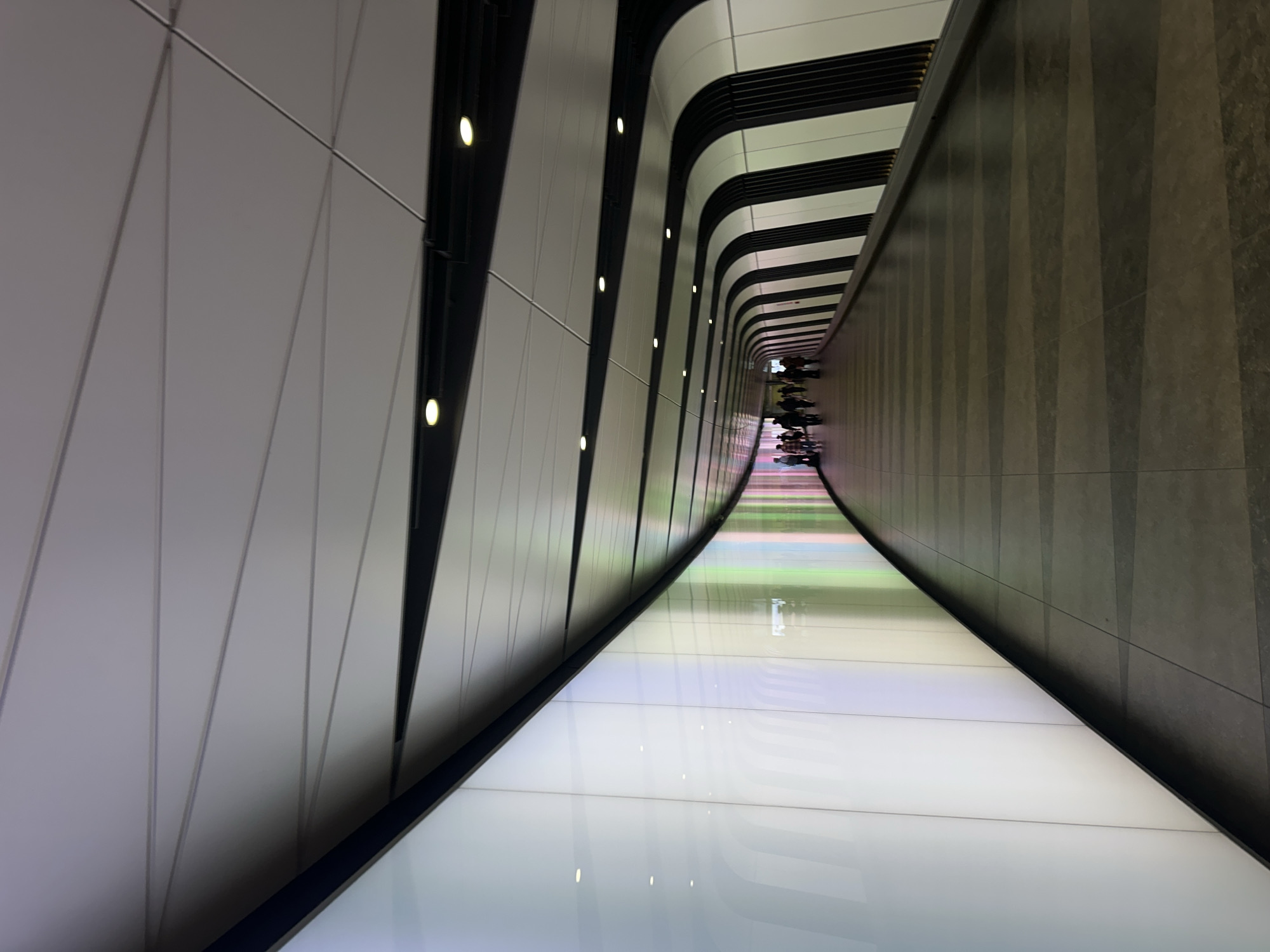 The image shows the pedestrian passageway in Terminal 5 of Heathrow Airport in London, United Kingdom. The corridor has an illuminated floor and curved walls with vertical stripes. People are walking in the distance.