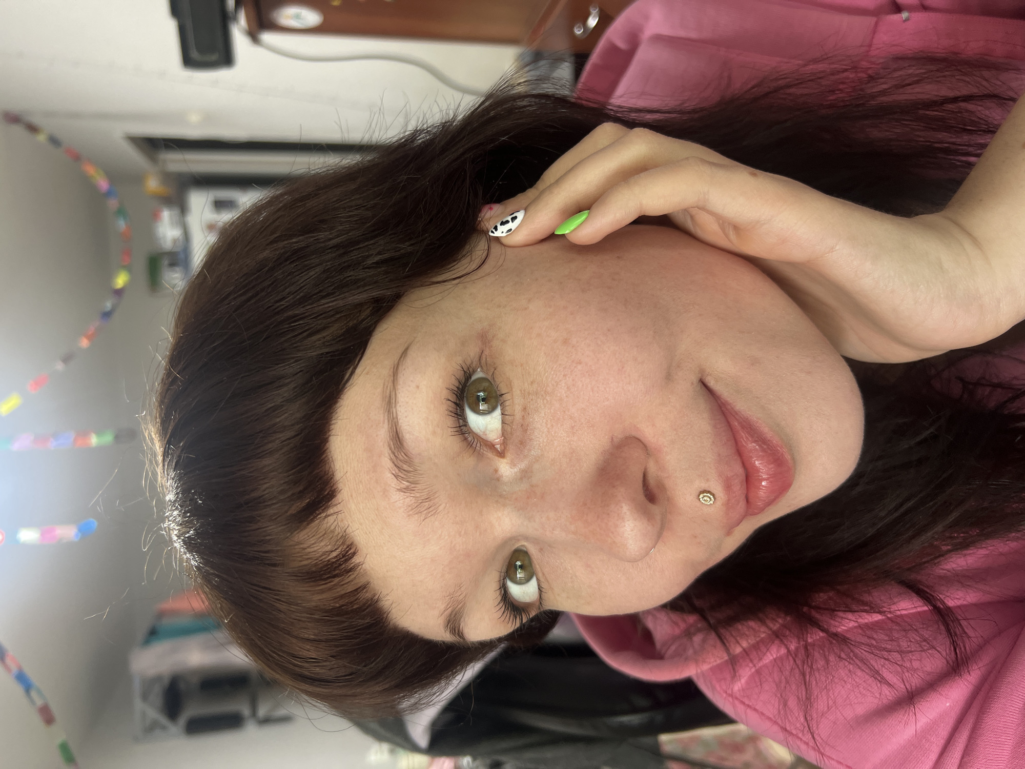 A young woman with fair skin, brown hair, and green eyes is the primary subject of the image. She has a lip piercing and her nails are painted with different patterns, including green and cow print. She is wearing a pink hoodie. The background is blurred, but suggests an interior setting. It is not possible to confirm the location as Chaikovsky, Russia based on the provided image alone.
