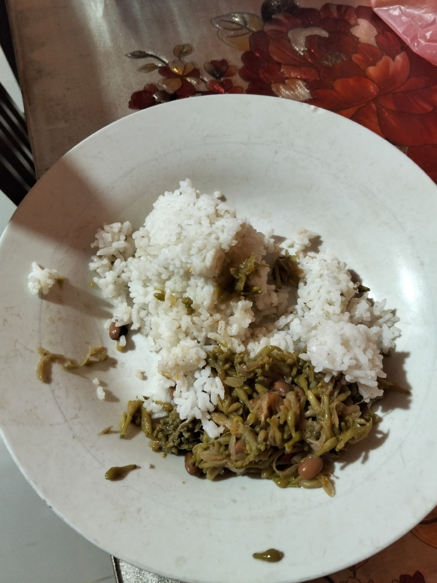 A close-up, overhead shot shows a white plate of food on a table. The plate contains white rice piled high on the left side and a mixture of cooked green vegetables and beans on the right. Some of the green vegetables appear to be shredded or finely chopped. A few stray beans and pieces of the green vegetable are scattered on the white surface of the plate. The table beneath is covered with a floral patterned tablecloth, featuring large red and orange flowers with brown and yellow accents. The lighting suggests an indoor setting, possibly a dining area in a home. The context of Payagaboh, Indonesia is not visually apparent from the image itself.