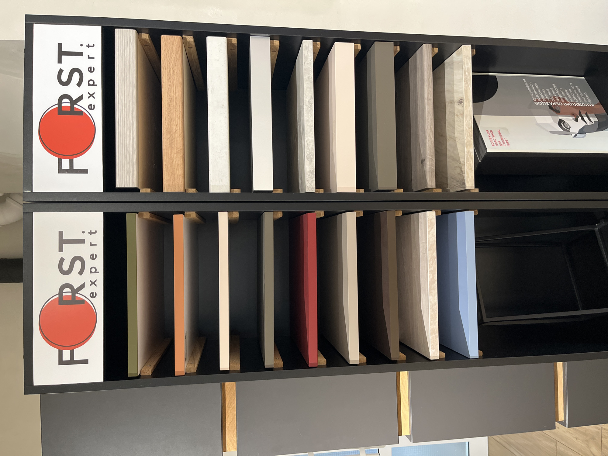 Here is a detailed description of the image:
The image displays a two-tiered display shelf, likely in a retail or showroom environment, showcasing various surface material samples, possibly for countertops or similar applications.
On the left side of each tier, there is a vertically oriented sign reading "FORST expert" with a large red circle integrated into the text. The shelves are filled with rectangular sample pieces in a range of colors and textures, including wood grains, solid colors like green, orange, red, and blue, as well as shades of gray and white. Each sample is placed vertically within the shelf dividers.
On the upper-right shelf, there is a white rectangular object. The object has text in a non-English script.
The shelves themselves are constructed from a dark material, possibly painted wood or metal. The dividers supporting the samples appear to be made of a lighter wood. Below the bottom shelf, glimpses of cabinetry in various shades of gray and light wood can be seen.
Overall, the image appears to be a display of surface material samples in a professional setting, designed to showcase variety in color, texture, and material.
