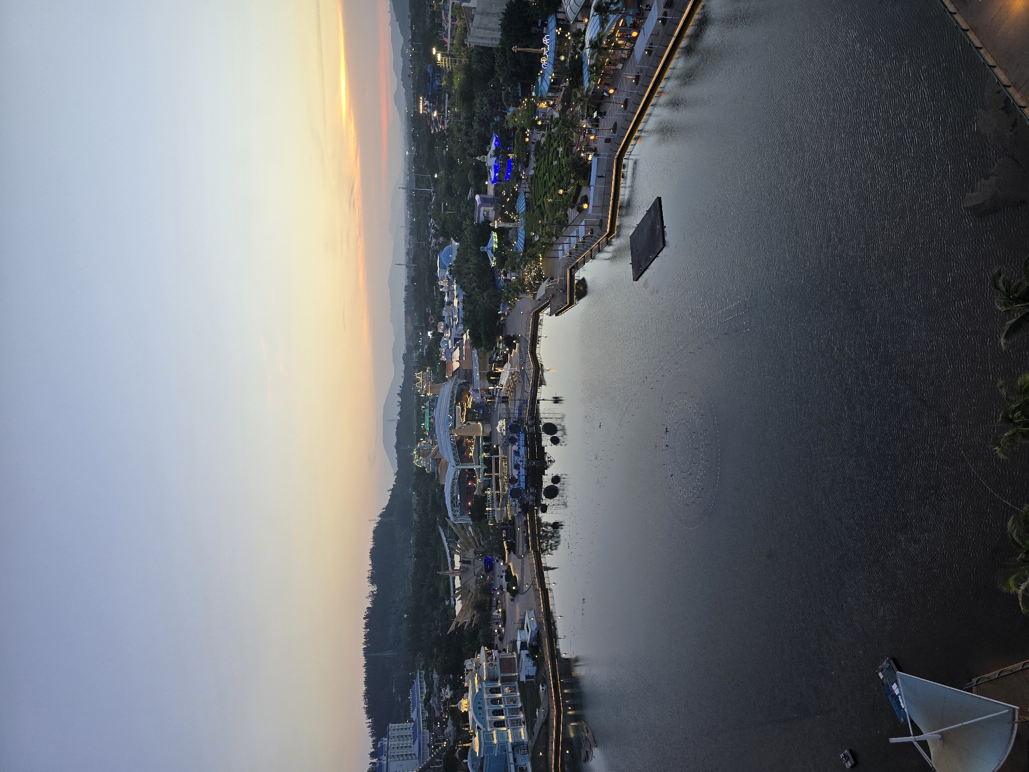 This is a high-angle, wide shot of a waterfront amusement park in Li'an, China, during twilight. The sky transitions from a pale blue at the top to a warm orange and yellow near the horizon, indicating either sunrise or sunset. The park features a large, dark body of water in the foreground, with the shore lined by a promenade illuminated with string lights.
On the left side of the water, a light blue, multi-story building with white trim is visible. Across the water, the amusement park complex is illuminated with colorful lights. Various attractions, including what appear to be roller coasters, a Ferris wheel, and stage structures, are present. Green trees and lush landscaping are interspersed throughout the park. In the distance, hills covered in trees form the backdrop, with power lines and distant structures visible.
Several small, dark objects, possibly boats or rafts, are scattered across the surface of the water. A rectangular floating platform is positioned near the center of the water. There are also clusters of small, light-colored dots in a circular formation on the water's surface, likely part of a fountain or lighting display. A few people are visible walking along the illuminated promenade. The overall scene depicts a vibrant, yet tranquil, amusement park setting as daylight fades.