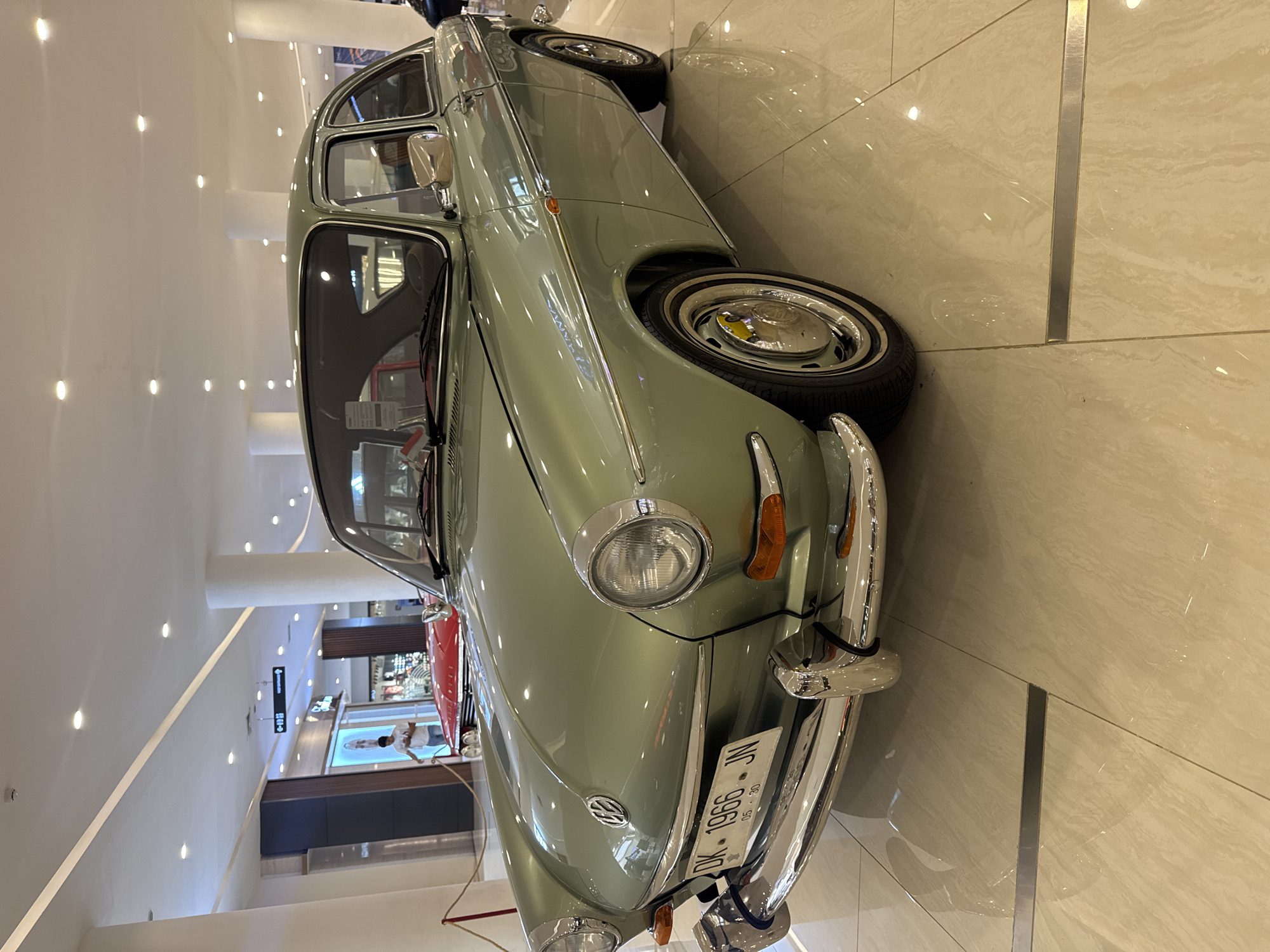 A light green Volkswagen Type 3 Notchback sedan, with license plate DK 1966 JN, is displayed on a light-colored tile floor in a shopping mall interior. The vehicle is oriented towards the viewer, with its front end in the foreground. A silver accent trim runs along the side of the car. The car is parked next to a tile floor section with a metal divider. Other stores and mall fixtures are visible in the background. Based on the license plate, the vehicle is located in Kuta, Indonesia.