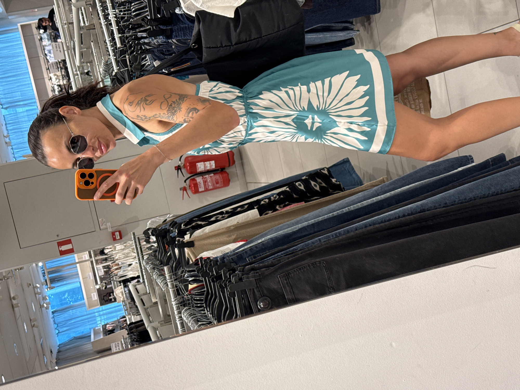 The image shows a woman taking a mirror selfie in what appears to be a clothing store. She is wearing a teal and white floral print dress, sunglasses, and has a ponytail. Her left arm is covered in tattoos. She is holding an orange-cased smartphone in her right hand. In the background, racks of clothes are visible, as well as fire extinguishers on the wall. The reflection in the mirror shows more of the store, including mannequins and shelving. There is no indication of the city of Pleven, Bulgaria, in the image or its context.
