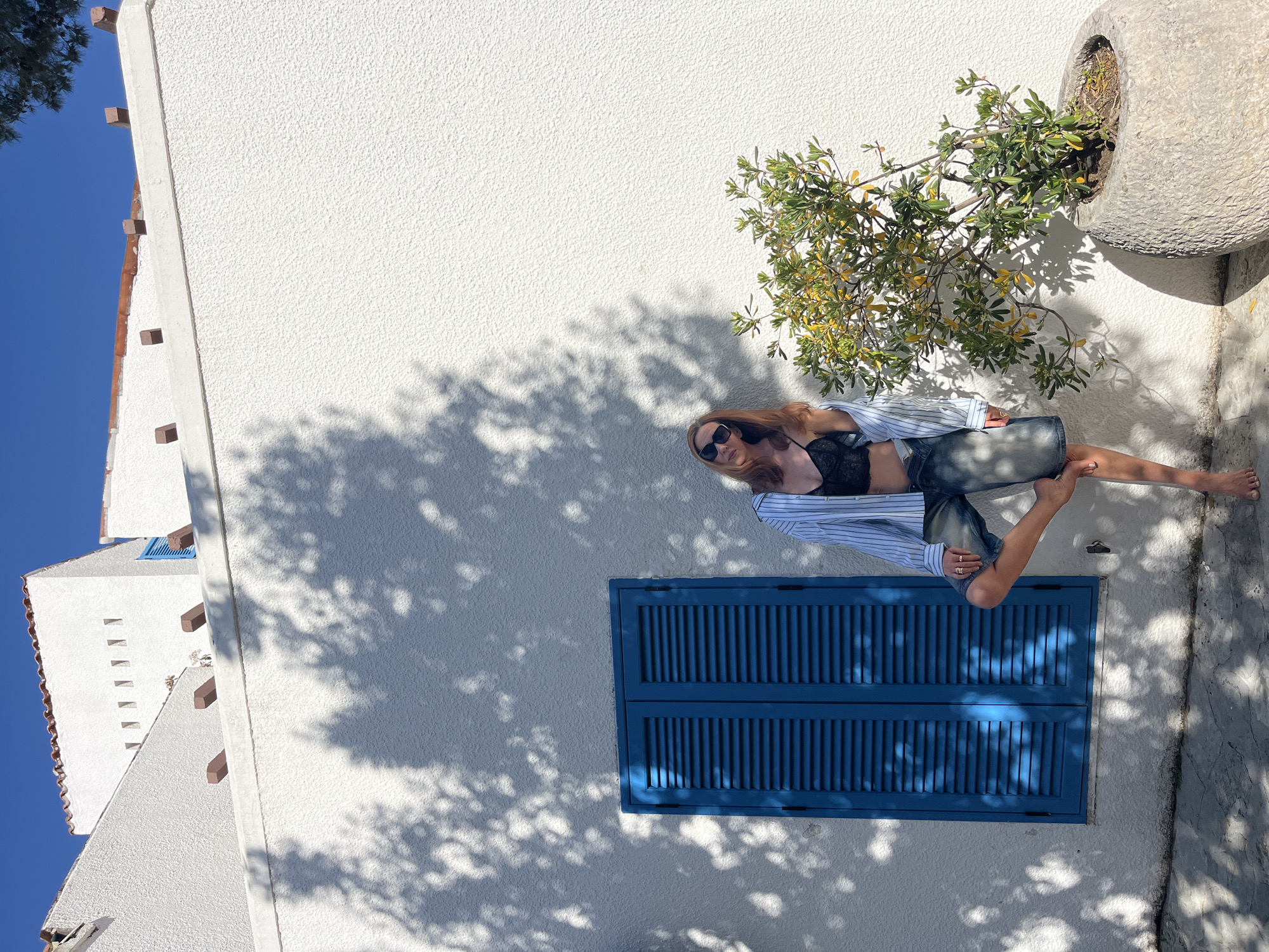The image captures a relaxed scene featuring a woman outdoors against a bright white stucco building in Budva, Montenegro. The woman, with light brown hair, wears sunglasses, a black lace bralette, and an open light blue and white striped button-up shirt over denim shorts or a skirt. She is lying on her back, propped up against the wall, with one leg bent and her bare feet visible.
Below her is a vibrant blue window with closed louvered shutters. To her right, a large, weathered grey stone planter holds a small green and yellow-leafed plant or shrub. The bright, clear blue sky is visible in the upper left, complementing the white architecture. Distinct, dappled shadows from an unseen tree are cast across the white wall and the woman, indicating strong, direct sunlight, suggesting it is mid-day.
The surrounding architecture includes other white buildings with visible terracotta roof tiles and decorative wooden beams under the eaves, reinforcing a Mediterranean or coastal European setting. The overall mood conveyed is one of leisure and sunshine. No text is visible in the image.