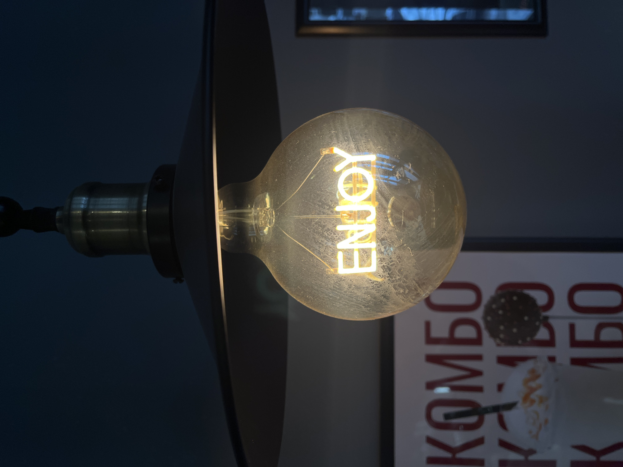 An illuminated lightbulb with the word "ENJOY" formed by its filament, hangs from a lamp fixture. The fixture is positioned in front of a wall with a poster visible in the background. The poster displays lettering in the Cyrillic alphabet. The lightbulb and the lamp fixture appear to be indoors, likely in a shop or restaurant in Krasnoyarsk, Russia.