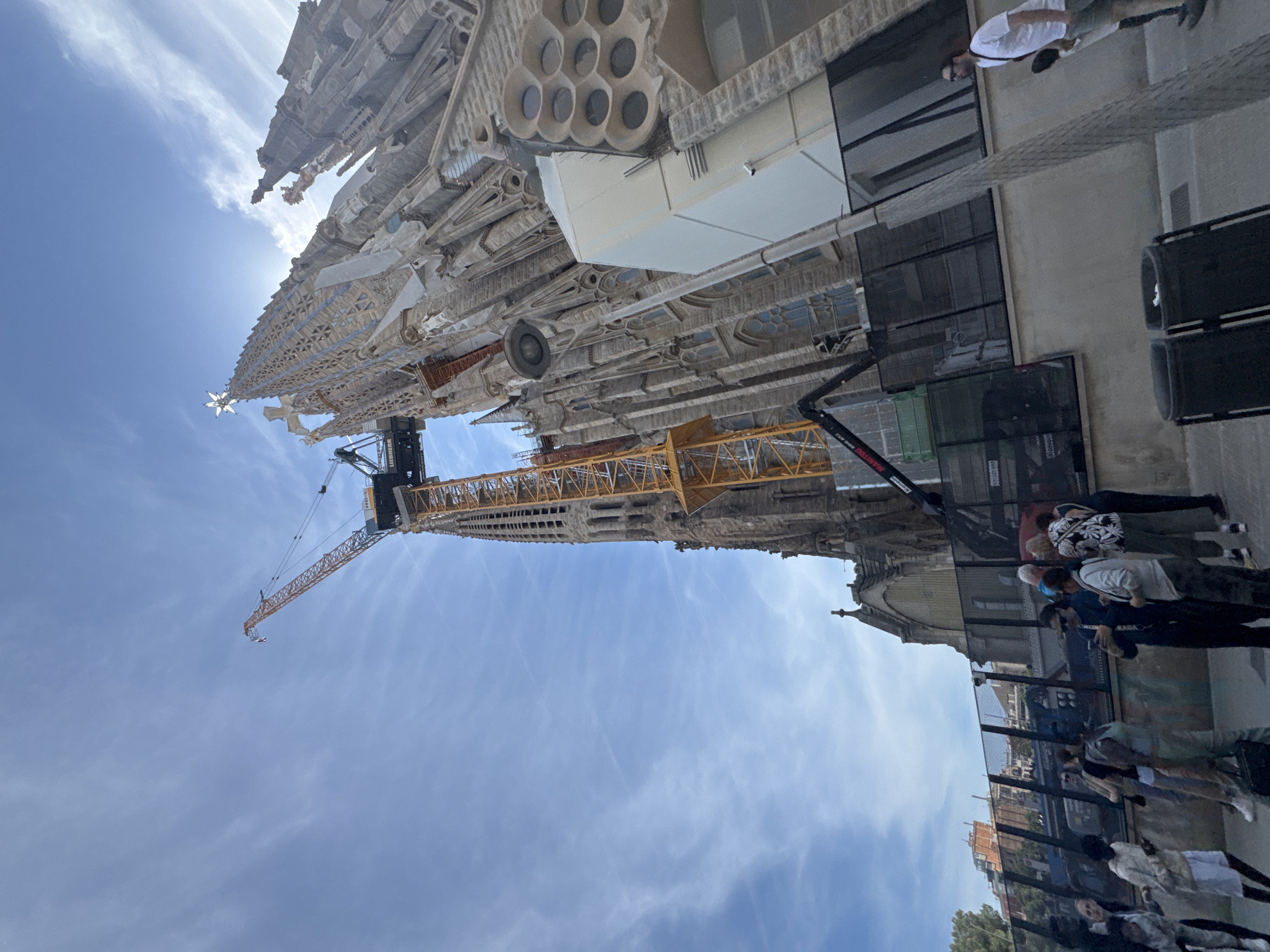 The image depicts the Sagrada Familia basilica in Barcelona, Spain, under construction. The basilica dominates the frame, revealing its intricate facade, spires, and a crane indicating ongoing construction. A portion of a building adjacent to the basilica is also visible. A group of people is standing near a fence. The sky is blue with scattered clouds.