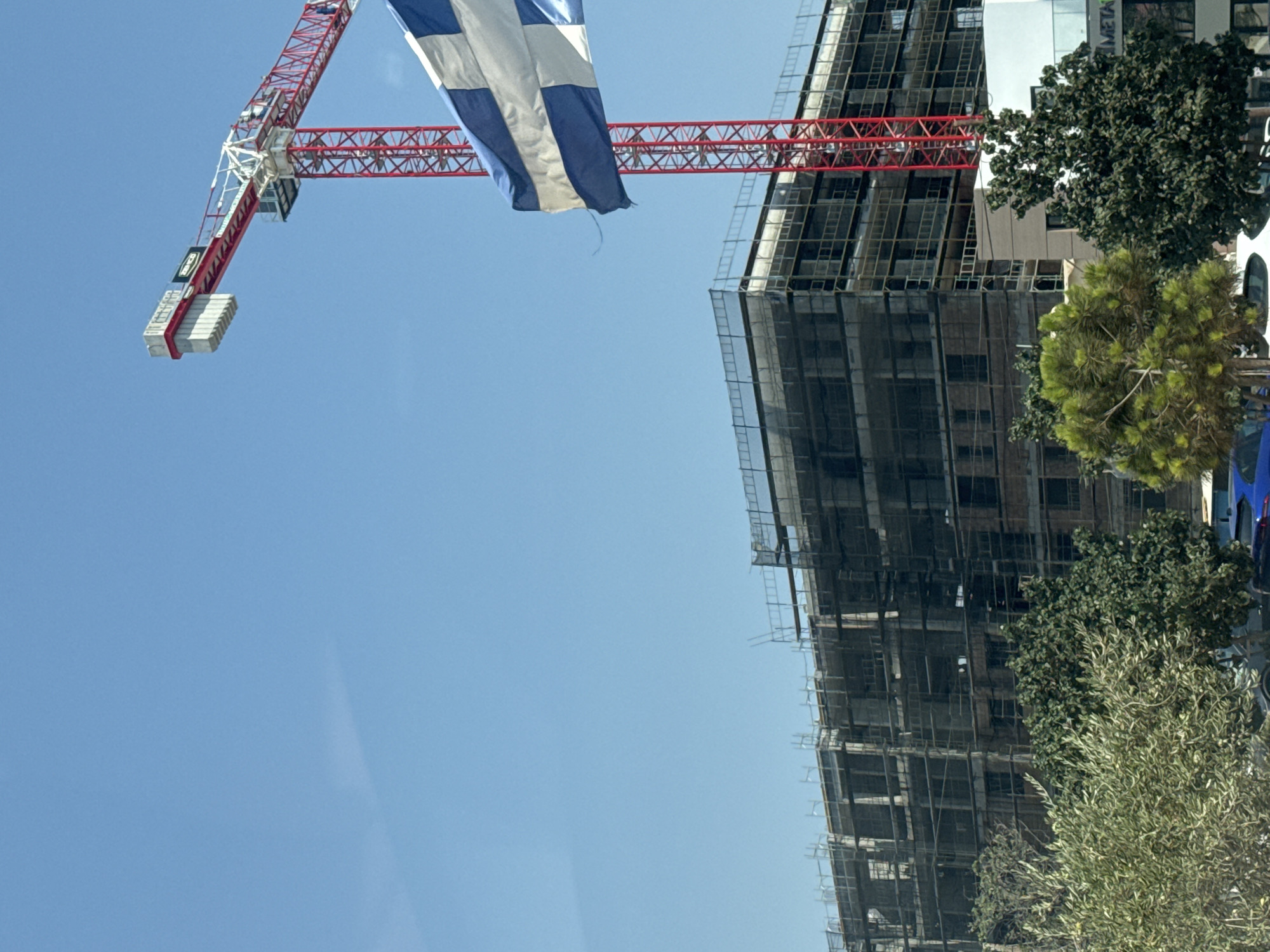 The image captures a construction site in Limassol, Cyprus, under a clear blue sky. A prominent red tower crane is visible, its jib extending to the right. A Greek flag, with its blue and white stripes, is unfurled, partially obscuring the crane. In the background, a multi-story building is under construction, covered in scaffolding and netting. The lower portion of the image features lush green trees, likely ornamental, framing the view of the construction. The scene suggests ongoing development and urban renewal within the city.