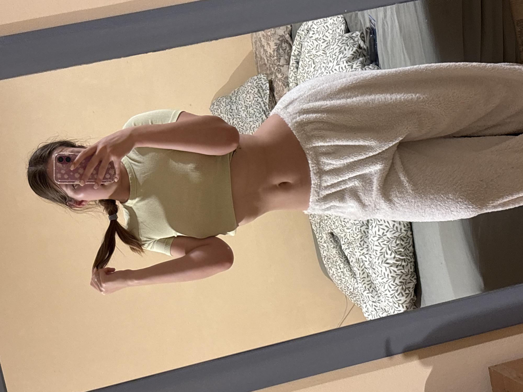 A female individual is visible in a mirror reflection. She is wearing a light green cropped top and white fuzzy sweatpants. The background appears to be a beige wall and a bed with patterned pillows and a light green comforter. The location of Bečići, Montenegro is not discernible from the image.