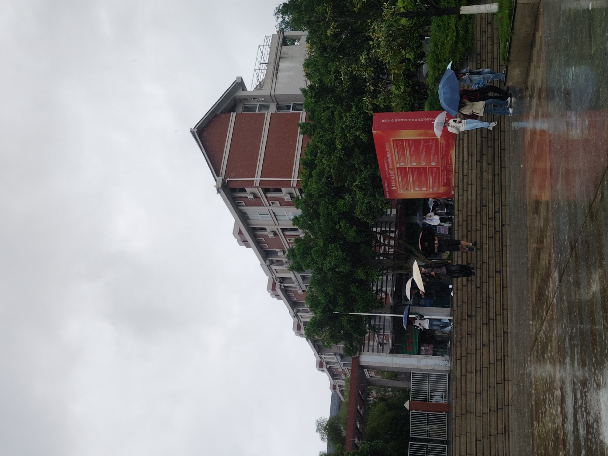 The image depicts an outdoor scene on a gray, overcast day, suggesting rain or recent rainfall, as evidenced by the wet, reflective ground and several individuals holding umbrellas. The setting appears to be a public or institutional area, consistent with the Chongqing High-tech Zone in China.
In the foreground and midground, multiple people are visible. One person, wearing a white jacket and light blue pants, holds a white umbrella while standing near a large, vertical red display board. Another individual nearby is seen holding a blue umbrella. Other people, some partially obscured, are walking on a brick-paved path or steps, also sheltering from the rain.
To the left, a multi-story building constructed of red brick with white window frames and trim extends into the background. Air conditioning units are mounted on the building's facade. Tall, green trees line the area between the building and the path. The sky above is uniformly cloudy and light gray.
A prominent feature is the large red informational display board. The text on the board is in Chinese characters, and the top section clearly states "中共中央 国务院印发" (Issued by the Central Committee of the Communist Party of China and the State Council). Below this title, the board contains multiple columns of text, likely detailing official policies, announcements, or guidelines, acting as a public information point. The overall scene conveys a typical rainy day atmosphere, with people going about their routines.