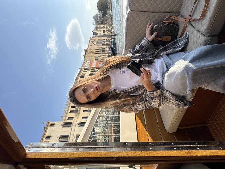 An adult female, appearing to be in her late 20s to early 30s, is seated in the passenger compartment of a wooden motorboat on the Grand Canal in Venice, Italy. She has long, straight brown hair and directly faces the camera with a neutral expression. Her attire consists of a white t-shirt with a dark graphic print, an open grey and black plaid flannel shirt, and light-wash blue jeans with a visible tear on the left thigh. On her left ring finger, a ring is present. She holds a black smartphone with a silver edge in her right hand, with her thumb on the screen. To her right, on the quilted beige seat, a brown Louis Vuitton monogram shoulder bag with a light leather strap is placed. The boat's interior features polished wooden panels and flooring. Across the sunlit Grand Canal, a series of multi-story historic Venetian buildings, or palazzi, display various earth tones including beige, yellow, and red. A contemporary building facade with large glass panels and a white awning is partially visible on the left bank. The sky is clear blue with a few isolated white clouds, indicating a sunny daytime environment.