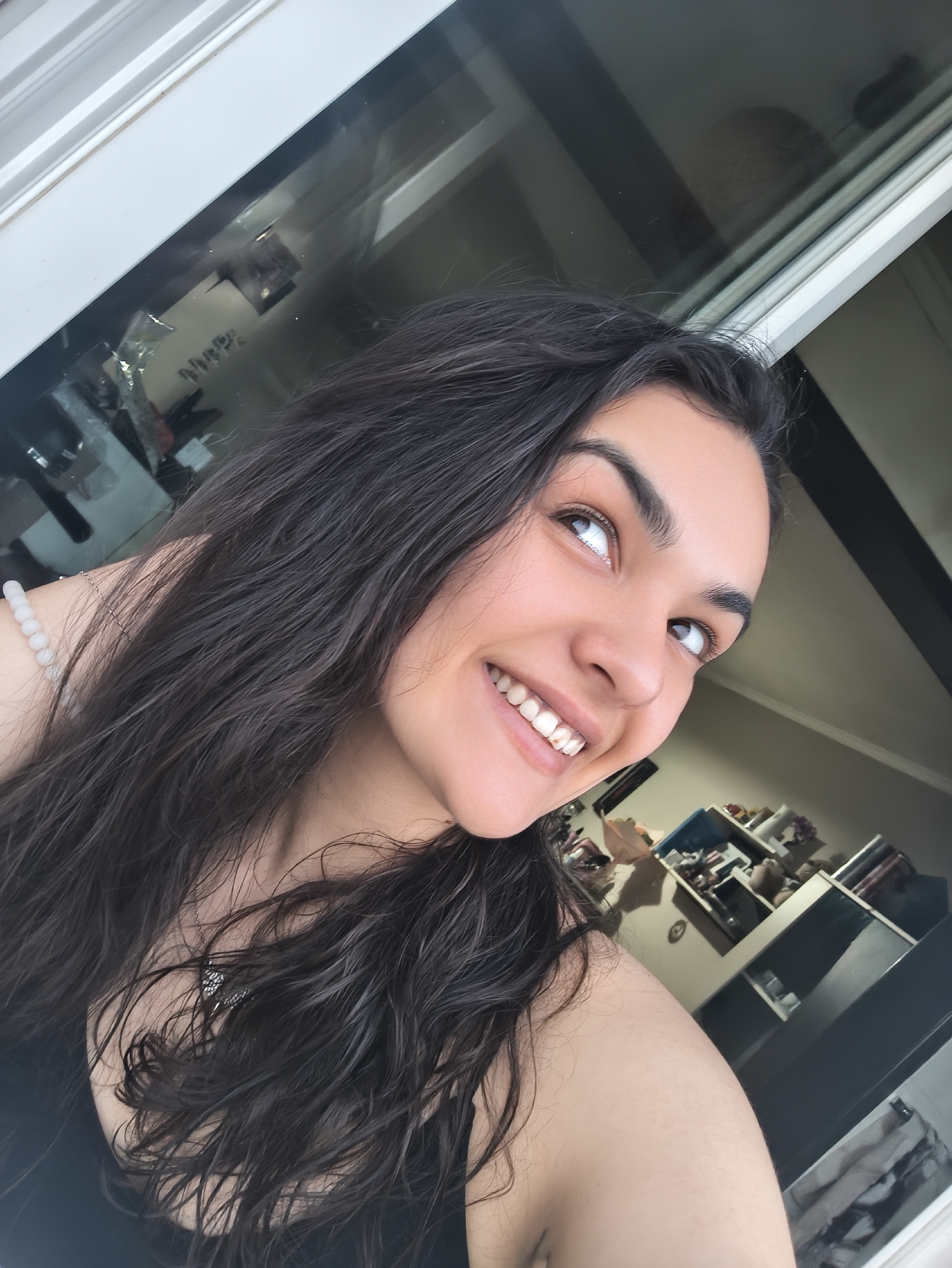A young woman with long, dark, wavy hair is captured in a close-up, angled selfie. She is smiling broadly, with her head tilted upwards and to the right, her eyes looking in the same direction. She appears to be indoors, with a window or glass door behind her that reflects a cluttered shelf unit. The lighting suggests it is daytime. The woman is wearing a black top and a beaded bracelet on her left wrist. The background, though out of focus, shows a room with shelves containing various items. There is no identifiable information that places this scene in Sofia, Bulgaria.