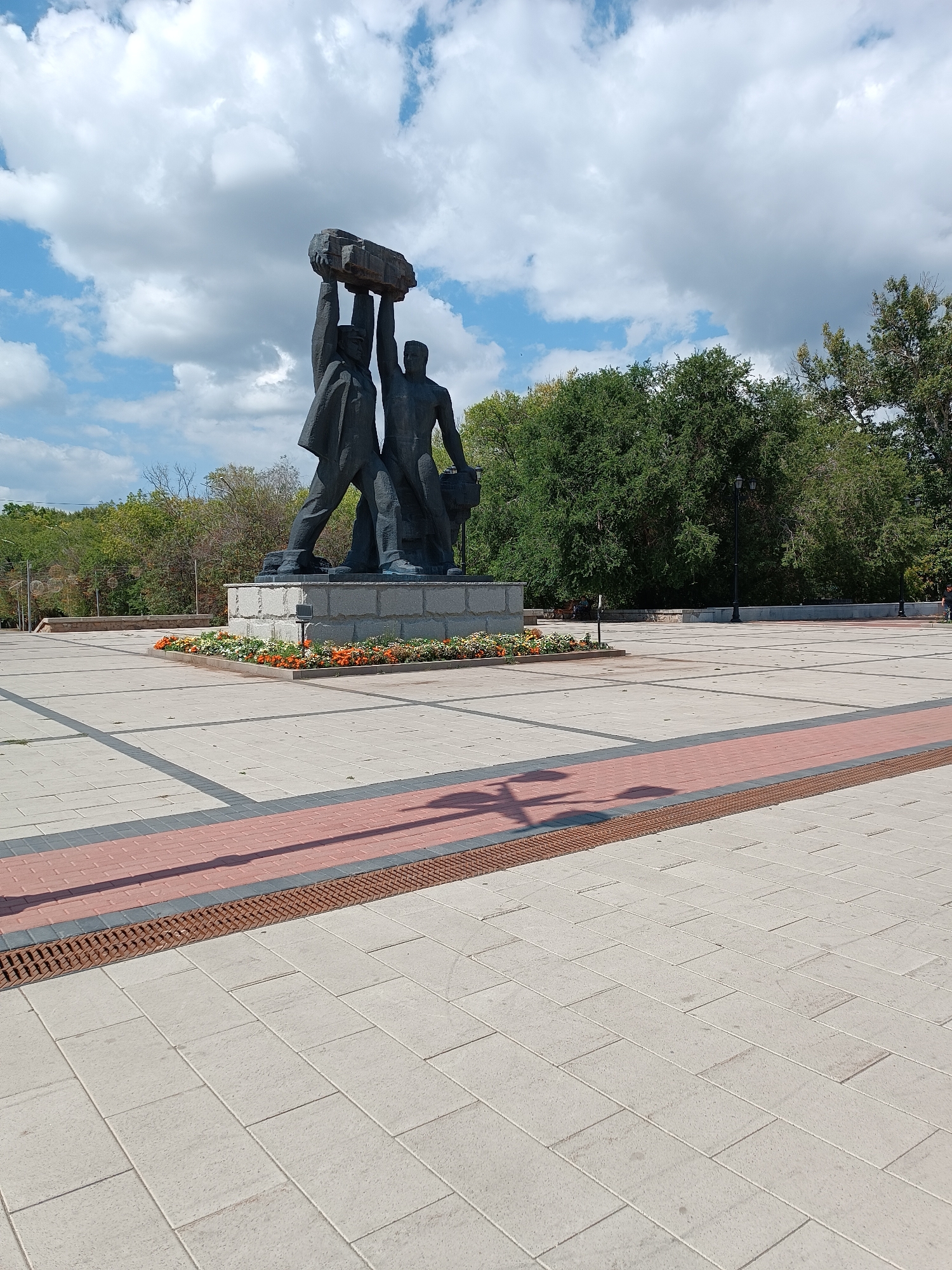 The image shows a bronze sculpture of two men holding a large stone above their heads, standing on a multi-tiered granite pedestal. The pedestal is surrounded by a flower bed with orange and white flowers. The sculpture is located in a large, paved public square. The pavement is made of light-colored square tiles with darker rectangular inlays forming a grid pattern. A red brick pathway runs diagonally across the square in the foreground, bordered by a grate. Behind the sculpture, a line of trees forms a backdrop, with a clear blue sky with fluffy white clouds above. A lamppost is visible to the right of the sculpture. The overall scene suggests an outdoor monument in a well-maintained urban space.