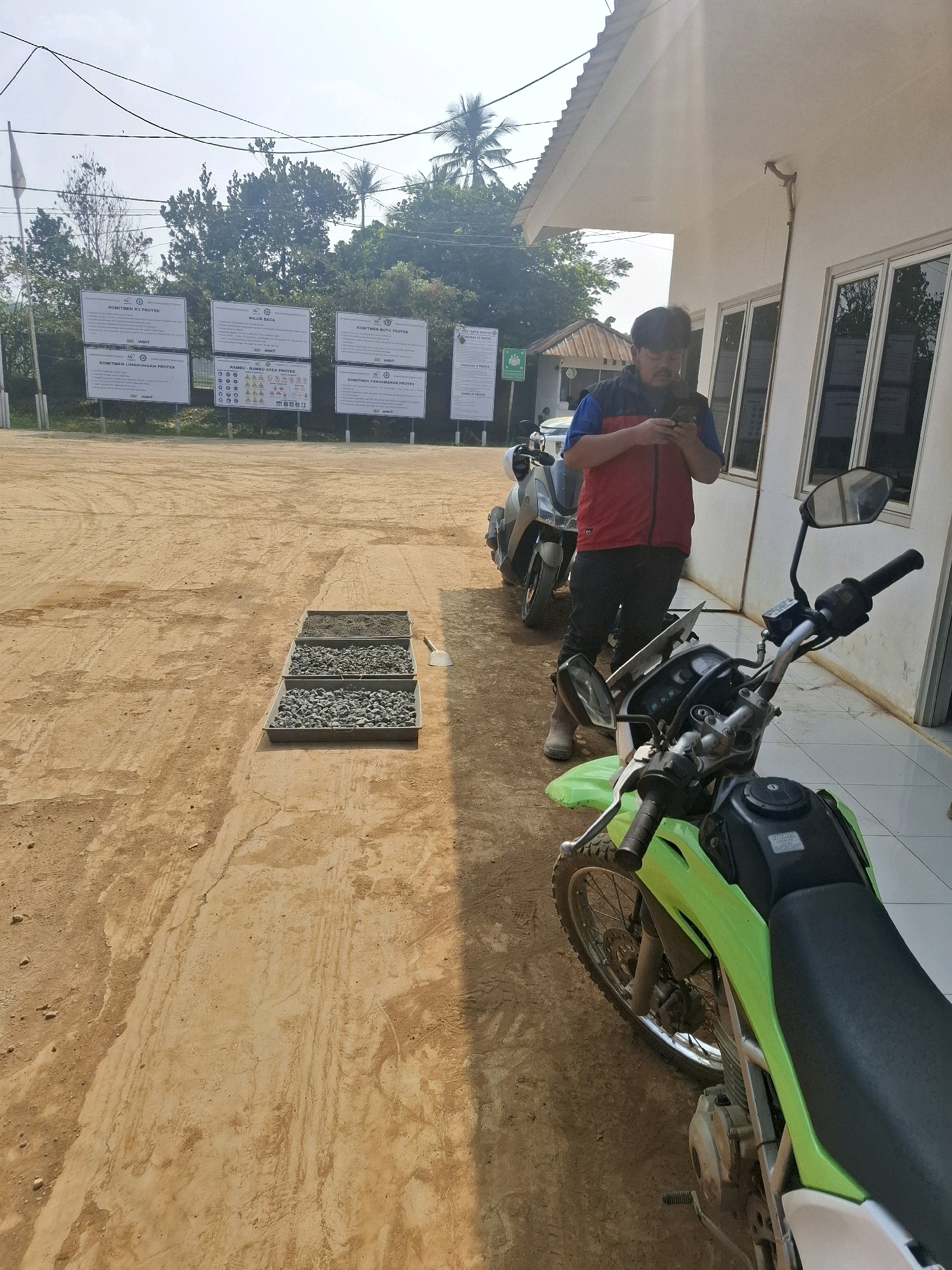 The image depicts an outdoor scene in Ciheulang Tonggoh, Indonesia. In the foreground, a bright green motorcycle is parked on a dirt ground. To the left of the motorcycle, three metal trays containing different types of gravel and sand are arranged in a row. In the background, a person wearing a red and blue vest over a dark shirt and dark pants is standing and looking at a mobile phone. Behind the person, two scooters are parked. Further in the background, a white building with several windows is visible, and several large informational billboards are mounted on posts. Trees and palm trees are also present, indicating a tropical or subtropical environment. The ground appears to be a mixture of dirt and sand, with some tire tracks visible. The sky is clear and the sun is casting shadows, suggesting it is daytime.