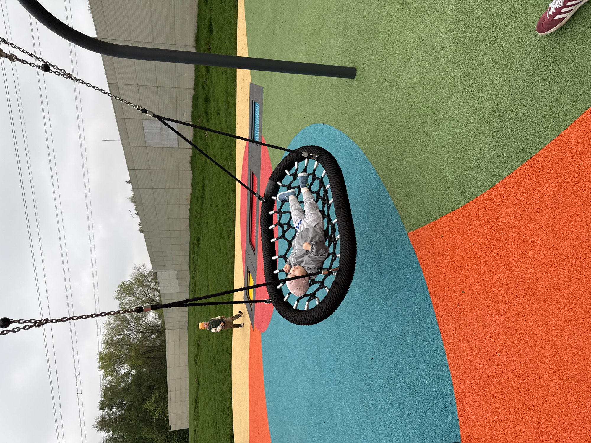A young child, wearing light-colored clothing and blue shoes, is lying face-up in a circular black net swing. The swing is suspended by chains from a metal frame, and it is located on a playground surface with sections of green, orange, blue, red, and yellow. Adjacent to the playground is a grassy area with trees, beyond which is a tall, gray barrier, potentially a sound wall. Power lines are visible in the background. A second child wearing brown clothing and a yellow hat is standing to the left of the swing. The location is possibly in Moscow, Russia.