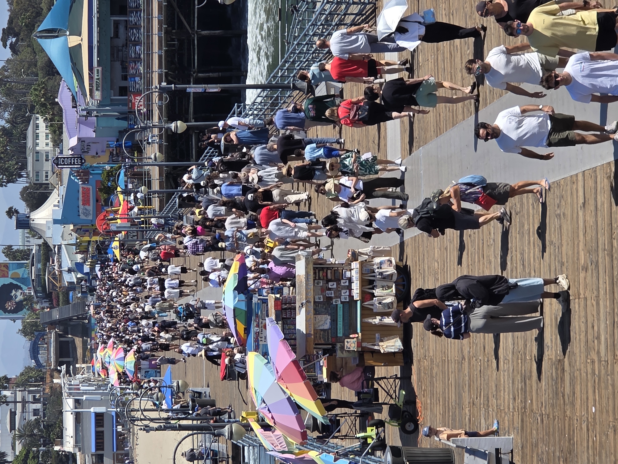 The image depicts a crowded wooden pier in Santa Monica, California. The pier is lined with various shops, food stalls, and umbrellas. Numerous people are walking along the pier, some are browsing stalls, and others are simply enjoying the atmosphere. The ocean is visible to the right of the pier, with waves crashing against the shore. The scene is bustling with activity, indicating a popular tourist destination. In the background, there are buildings and palm trees, characteristic of the Santa Monica coastline. The overall impression is one of a lively and vibrant pier on a sunny day.