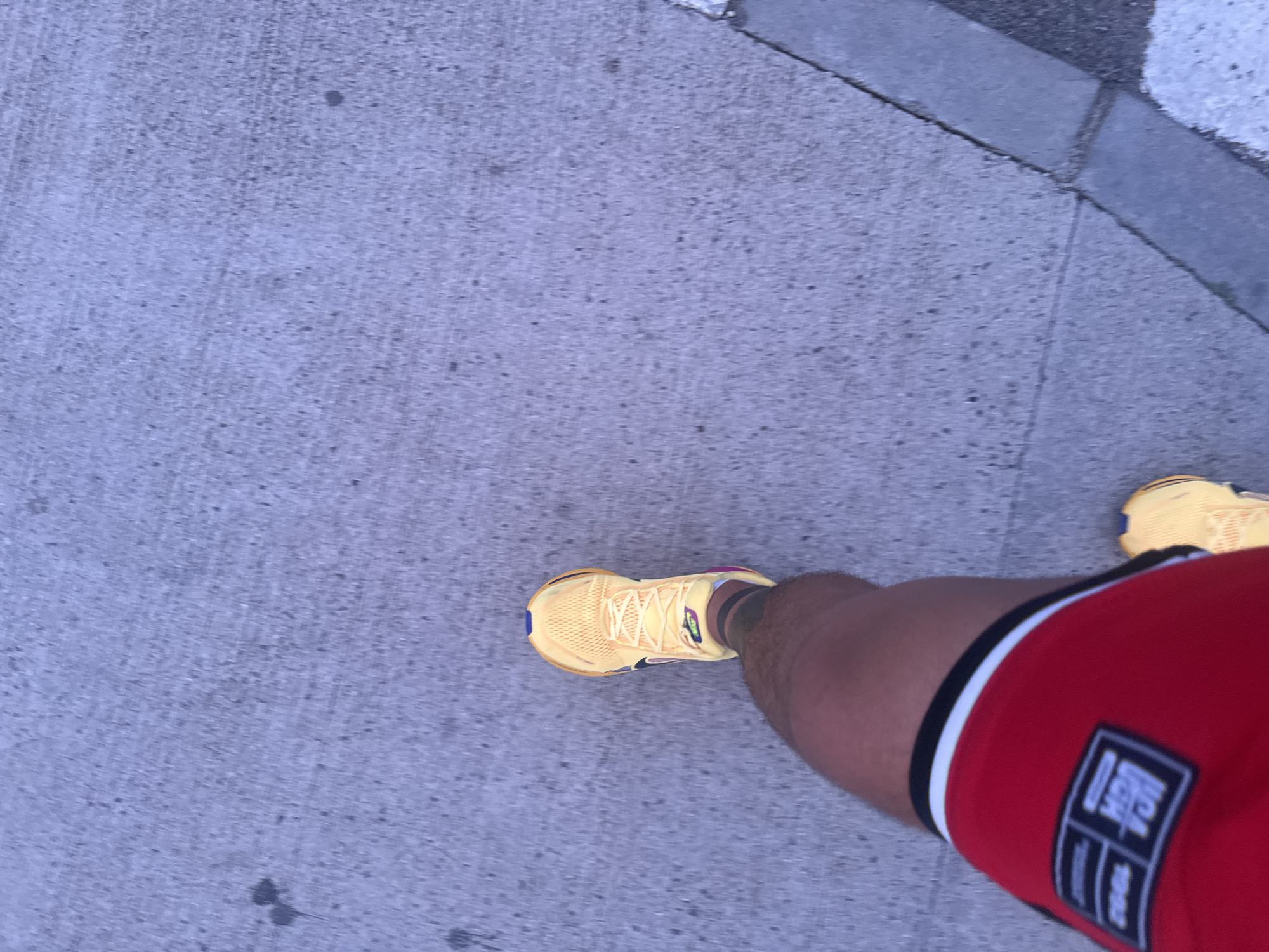 A low-angle, full-body shot shows a person walking on a concrete sidewalk in Boreti, Montenegro. The person is wearing bright yellow running shoes and red athletic shorts with a black and white stripe. The concrete sidewalk has visible cracks and is lightly speckled with dark spots. A gray curb runs along the right side of the sidewalk, separating it from a darker paved area. The lighting suggests it is dusk or dawn.