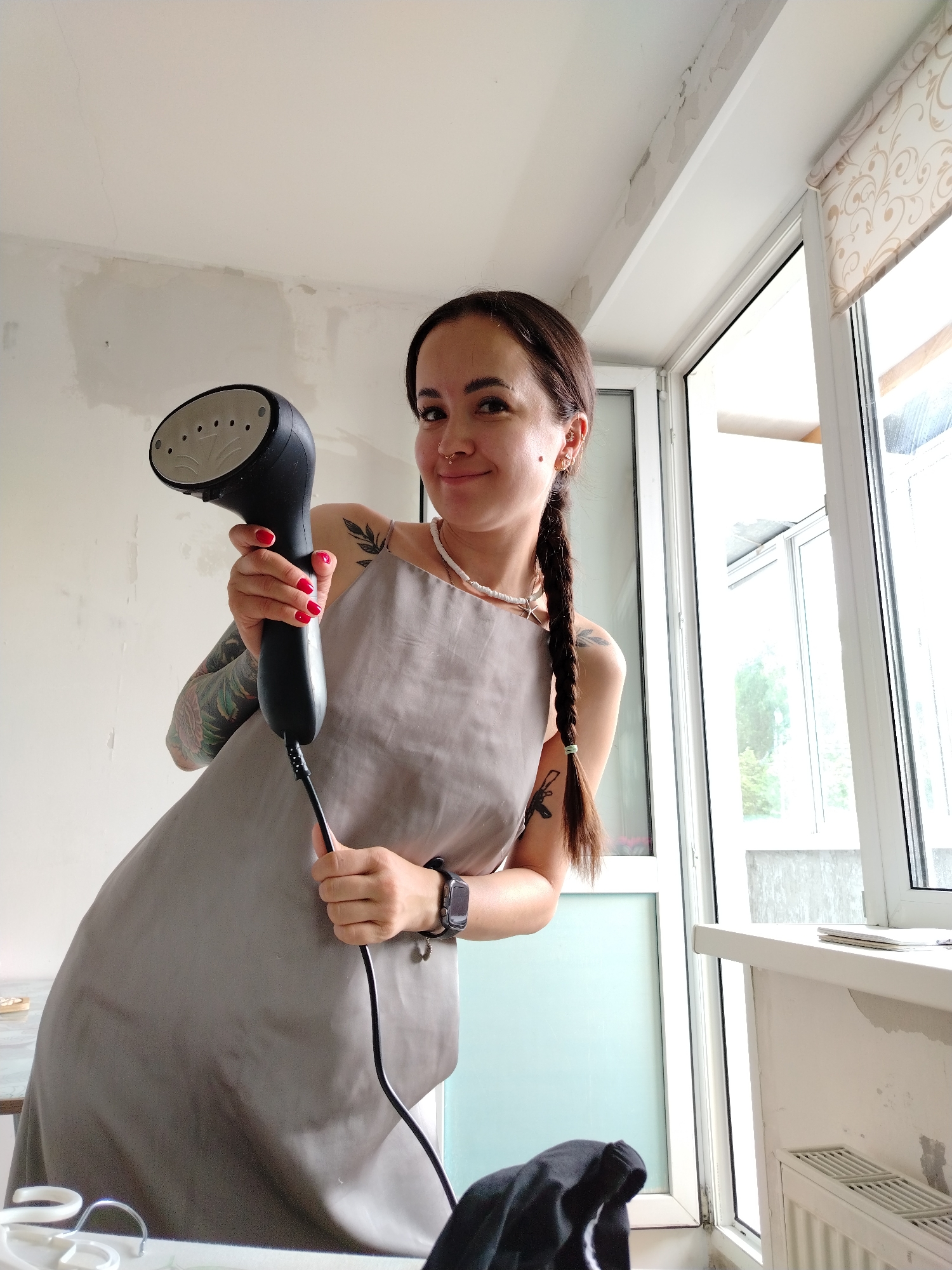 A young woman with dark hair in braids stands indoors, holding a black garment steamer. She wears a gray apron and has tattoos on her arms. The room appears to be under renovation, with exposed plaster on the walls. A window with blinds is visible on the right side of the frame, and part of a white radiator is visible below the windowsill. A table with some white objects, possibly hangers, is in the foreground. The overall lighting suggests it is daytime. The location is identified as Noviy, Russia.