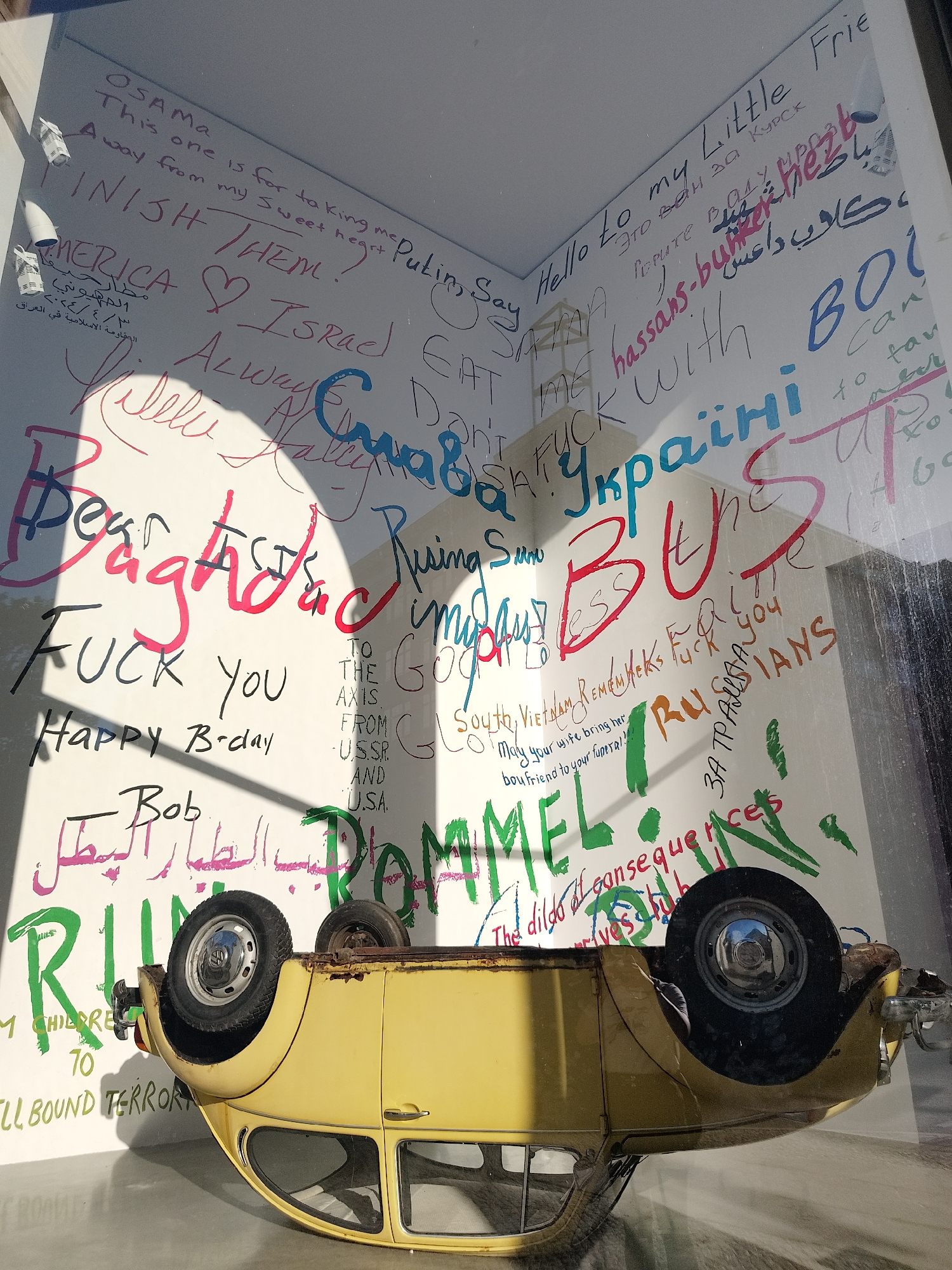 A yellow Volkswagen Beetle is overturned in what appears to be an indoor space. The surrounding walls are covered in graffiti in multiple languages, including English, Arabic, and Cyrillic. Phrases and words such as "OSAMA," "Putin," "FUCK YOU," "BAGHDAD," "ROMMEL," "Ukraine," "Israel," and "TERROR" are visible. The scene suggests a display or installation, possibly of a political or anti-war nature.