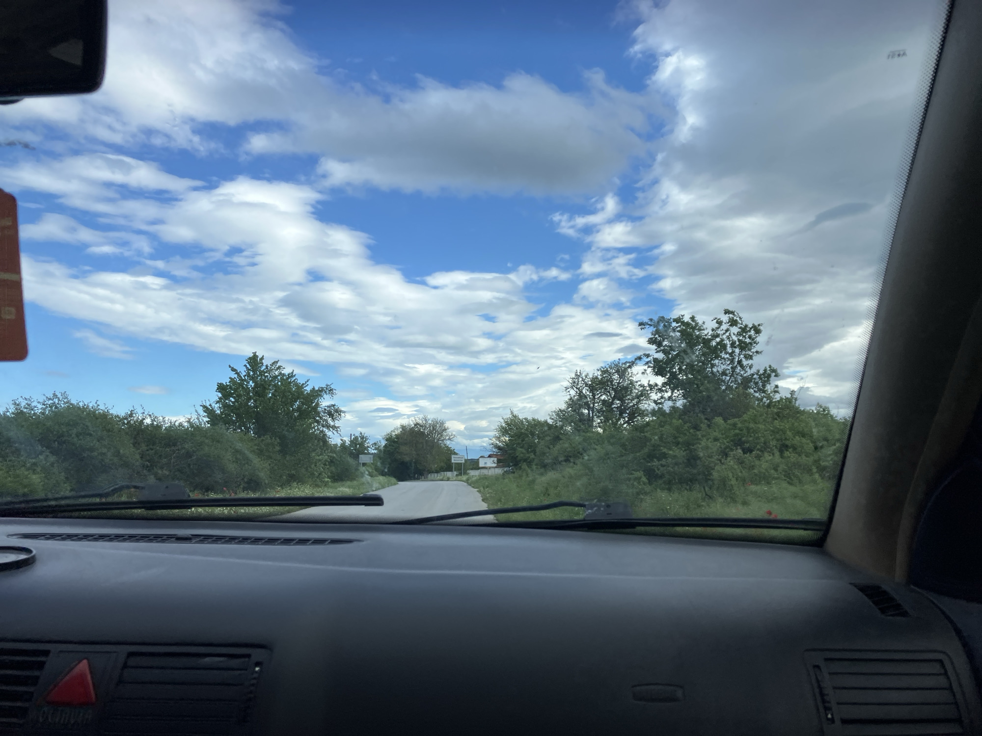 The image shows a view from inside a car driving on a road toward the village of Krasnovo, Bulgaria. The sky is partly cloudy with patches of blue. Vegetation, consisting of trees and bushes, lines both sides of the road. The car's dashboard and windshield wipers are visible. A small road sign is visible just beyond the trees in the distance.