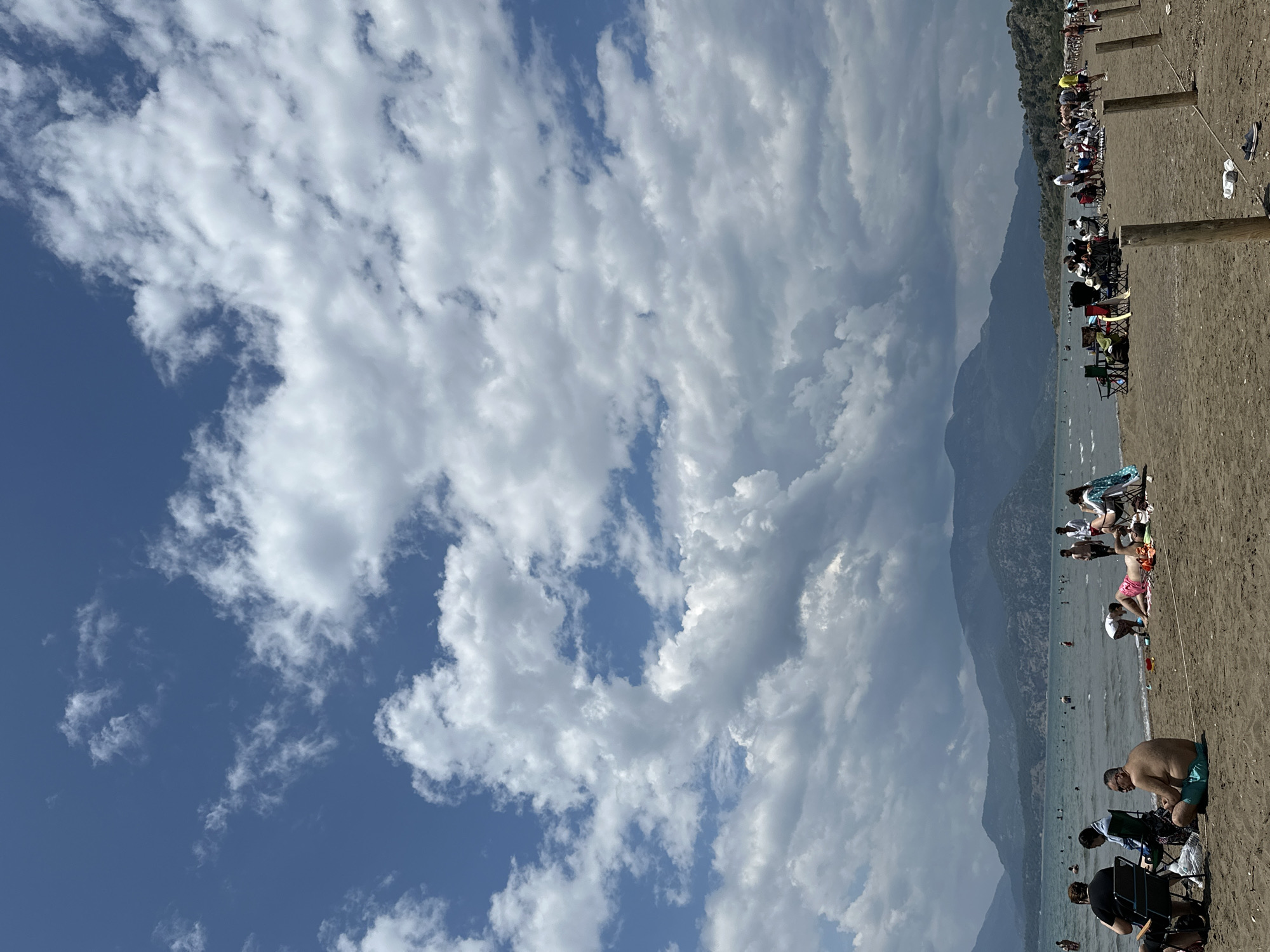 The image shows a beach scene in Ortaca, Turkey. The sky is predominantly blue with scattered white cumulus clouds. The beach is crowded with people sitting in beach chairs, swimming in the water, and walking along the shoreline. In the background, there are mountains covered in trees. The sand appears to be brown. The scene suggests a sunny day with a mix of open sky and cloud cover.