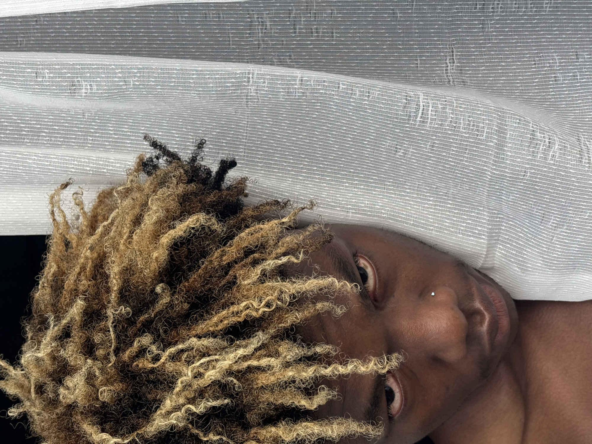 A close-up portrait features a person with dark skin, positioned at an angle against a light, textured fabric background. The person's head is tilted, with their gaze directed towards the viewer. Their hair is a prominent feature, consisting of voluminous, tightly coiled or loc'd strands in a blend of dark brown/black and lighter, possibly bleached, blonde or light brown tones.
Part of the person's face is visible, including both eyes, a portion of the nose featuring a small, shiny stud piercing on the nostril, and the lips. Their expression is neutral and direct. A hint of a bare shoulder or upper chest can be seen at the bottom right of the frame.
The background is a soft, light gray or off-white fabric with a distinct horizontal striped and slightly crinkled texture, possibly a curtain or bedding, draped to create gentle folds. The lighting is soft, highlighting the textures of the hair and fabric. No specific activity is depicted other than posing for the photograph. There is no visible text. The image was taken in Igboefon, Nigeria.