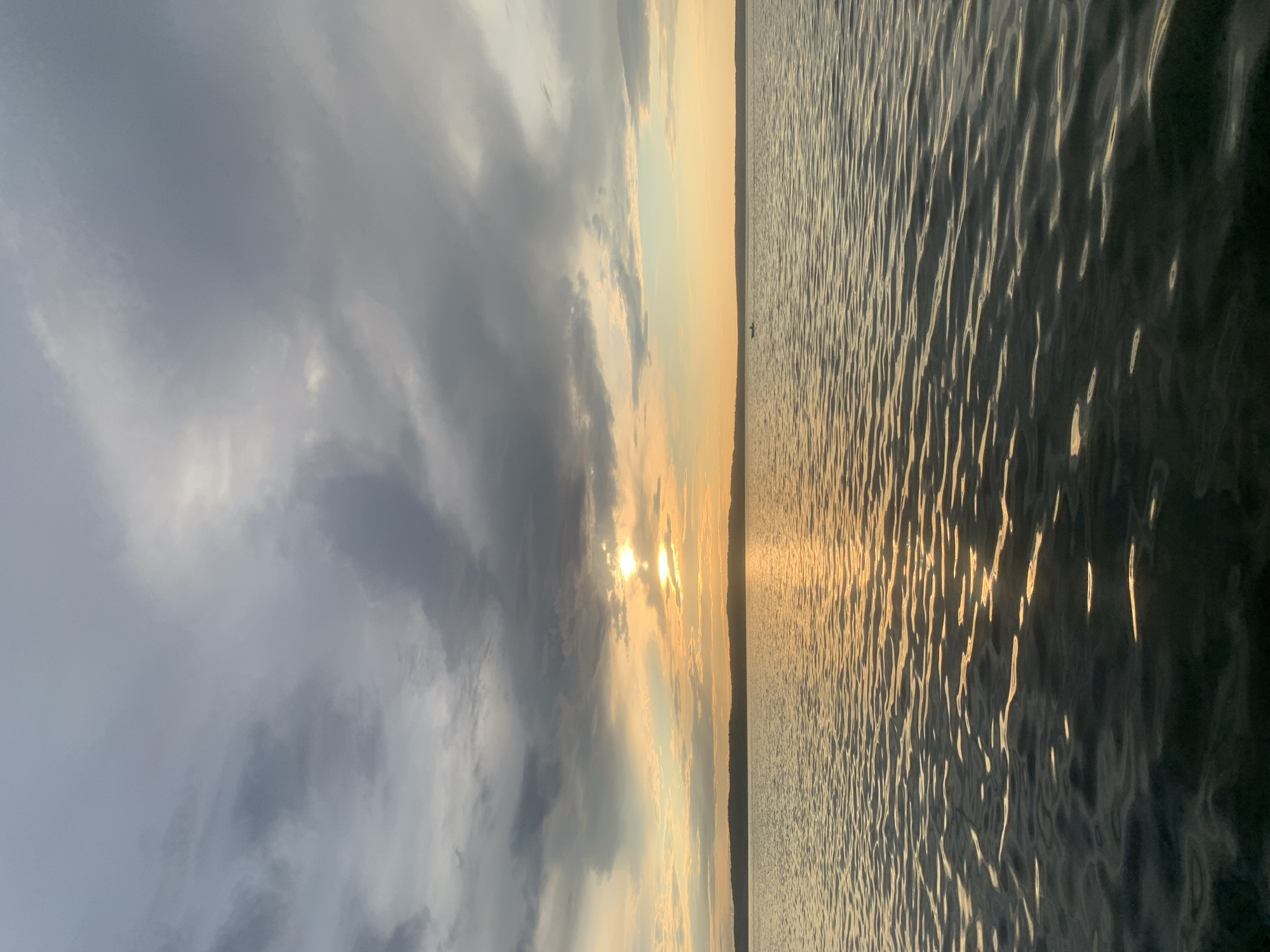 A calm body of water reflects the sunset and a cloudy sky. A thin, dark line of trees forms the horizon, with a small boat visible in the distance. The water is dark with ripples, and the sky is a mix of dark gray clouds and bright orange and yellow hues from the setting sun. The overall mood is serene and peaceful.