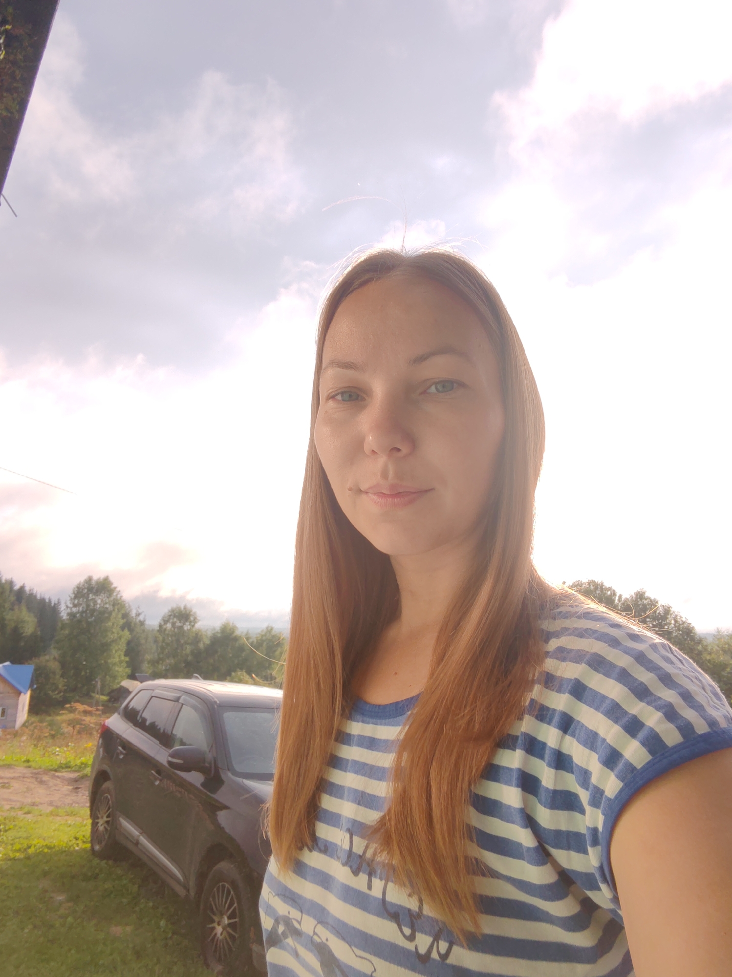 In a rustic outdoor setting, a woman with shoulder-length, reddish-brown hair, wearing a blue and white striped t-shirt, poses for a selfie. She is positioned in the foreground, with a slightly overcast sky providing a bright backdrop. To her left, a dark-colored SUV is parked on a grassy area, with a small, blue-roofed structure visible in the distance, suggesting a rural or semi-rural environment. The surrounding area is dotted with trees, indicating a natural or undeveloped landscape. The overall impression is a casual snapshot taken during daylight hours.