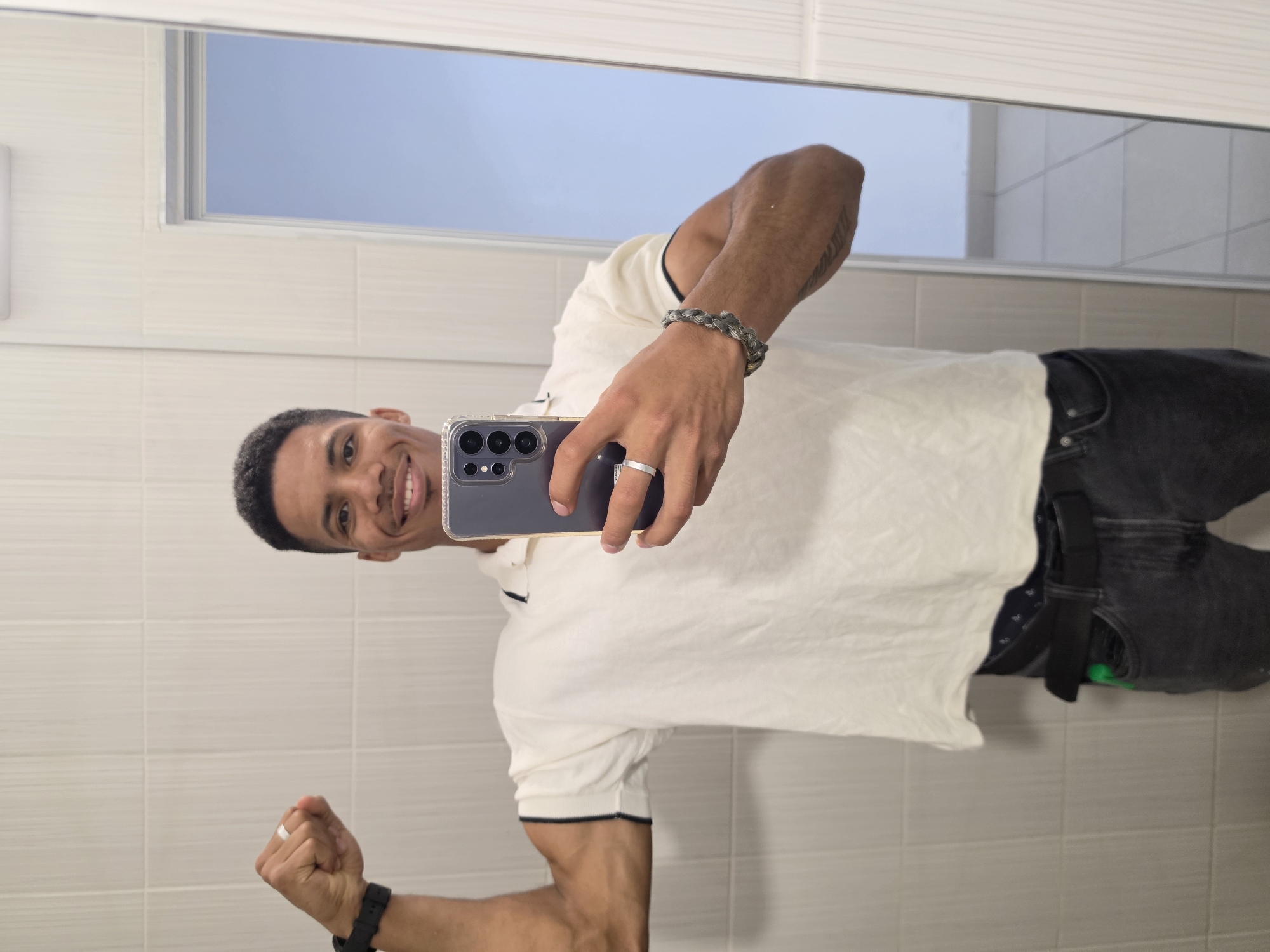 The image shows a young man taking a mirror selfie in what appears to be a tiled bathroom. He is wearing a white polo shirt with dark trim, dark grey jeans, and a black belt. He is flexing his left bicep and holding a smartphone in his right hand. He has a white ring on his left hand and a beaded bracelet on his right wrist. The background consists of white tiled walls and a partial reflection of a blue sky. There is no indication of the city of Varna, Bulgaria, or any specific urban location.