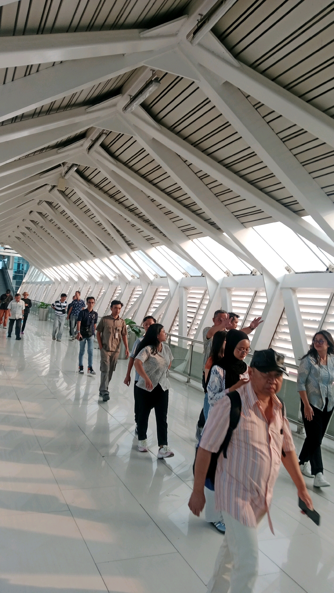 This is a medium shot, eye-level, exterior vertical photograph of people walking down a modern, covered pedestrian walkway, likely at an airport or transit hub, in Tangerang, Indonesia. The walkway features a white, angular, skeletal roof structure with angled skylights, allowing natural light to illuminate the glossy white floor. The floor reflects the people walking on it, creating elongated shadows. The people are of various ages and ethnicities, dressed in casual and professional attire, some carrying bags. Most are walking away from the camera, forming a line. One man in the foreground, wearing a pink and white striped short-sleeved shirt and white pants, is walking towards the camera and holding a dark rectangular object in his right hand. A woman in a light blue patterned top and black pants is walking towards the camera on the far right. In the middle ground, a man in a brown uniform shirt and khaki pants is walking away from the camera, and a woman in a white patterned blouse and black pants is also walking away. The overall impression is one of orderly movement through a contemporary architectural space.