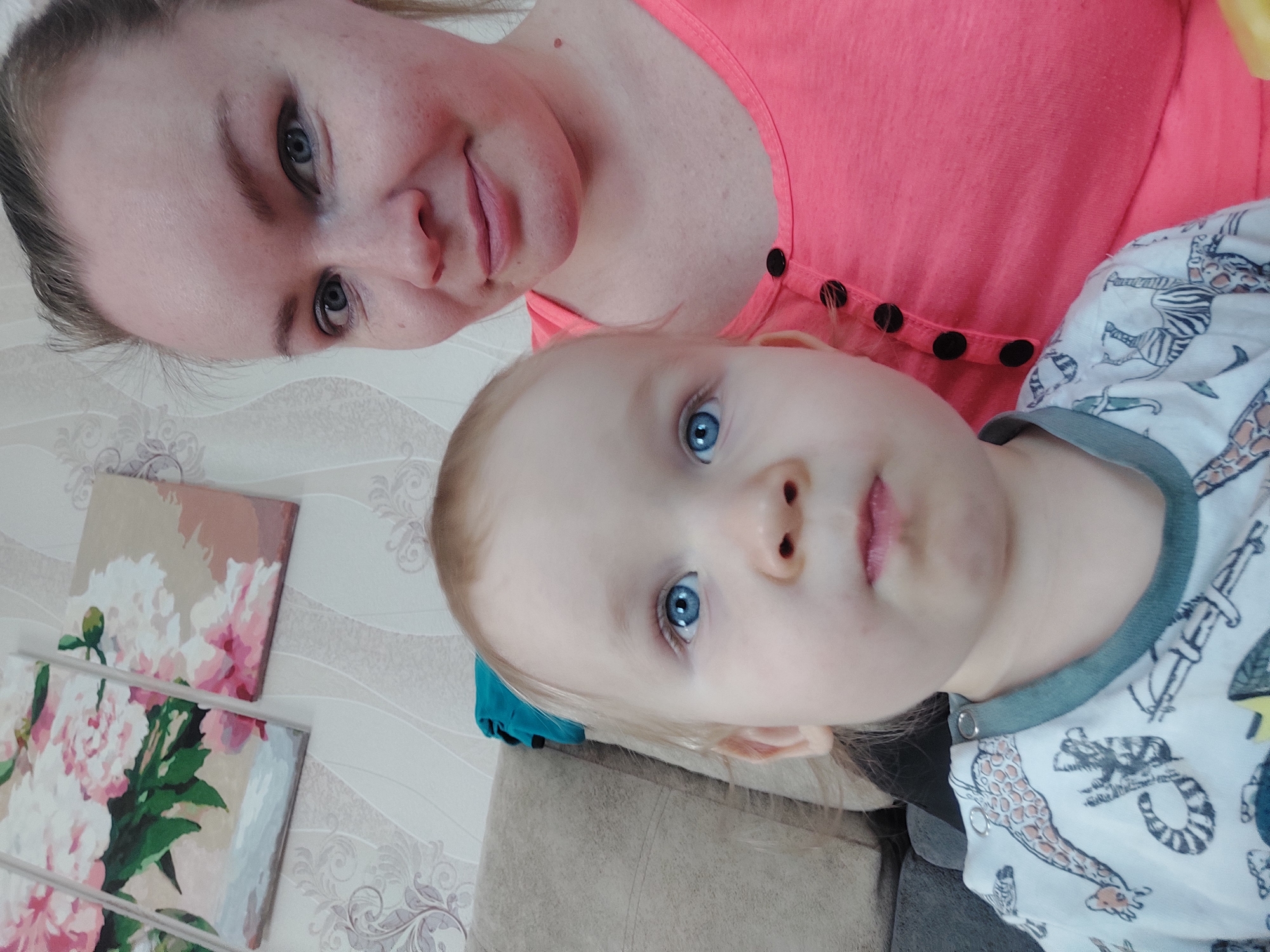 A mother and child are posing for a selfie, turned on its side. The mother has light skin, blue eyes, and blonde hair pulled back. She is wearing a pink shirt with black buttons. The child is light-skinned with blue eyes and light blonde hair. They are wearing a white shirt with a grey collar and a cartoon animal print. Two paintings of pink flowers are on the wall behind them. They are in what appears to be a home, likely in Большой Букор, Russia.