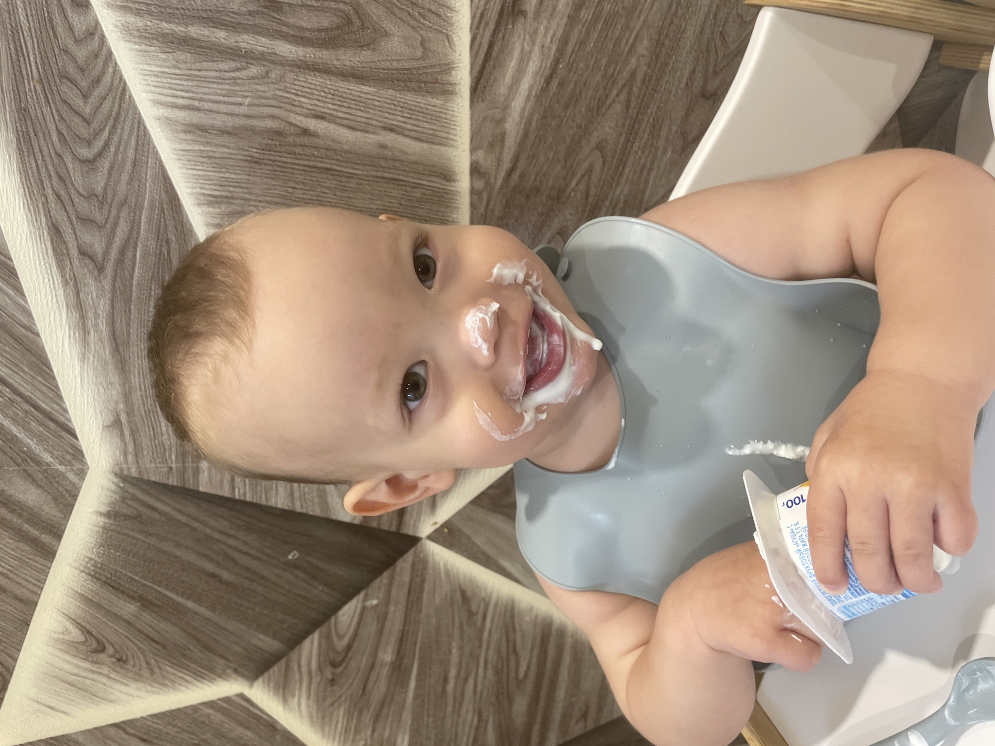 A baby is sitting in a high chair, covered in yogurt. The baby is wearing a grey bib and holding a yogurt container. The background appears to be a room with patterned flooring and part of the high chair. The container indicates that the photo was likely taken in Russia. I cannot definitively determine if the image was taken in Perm, Russia, with only the provided evidence.