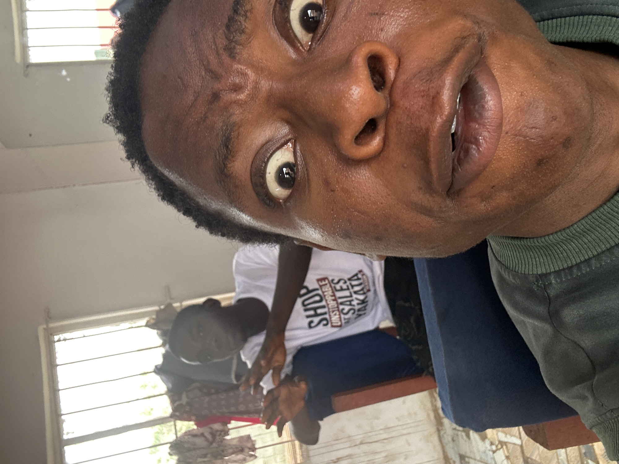 A selfie shows a Black man with a dark green sweatshirt. The man's face is close to the camera and his eyes are wide. Another man is seated in the background. He is wearing a white T-shirt with the word "SHOP" printed on it. The location is a room with a window with blinds. The city and location are unknown.