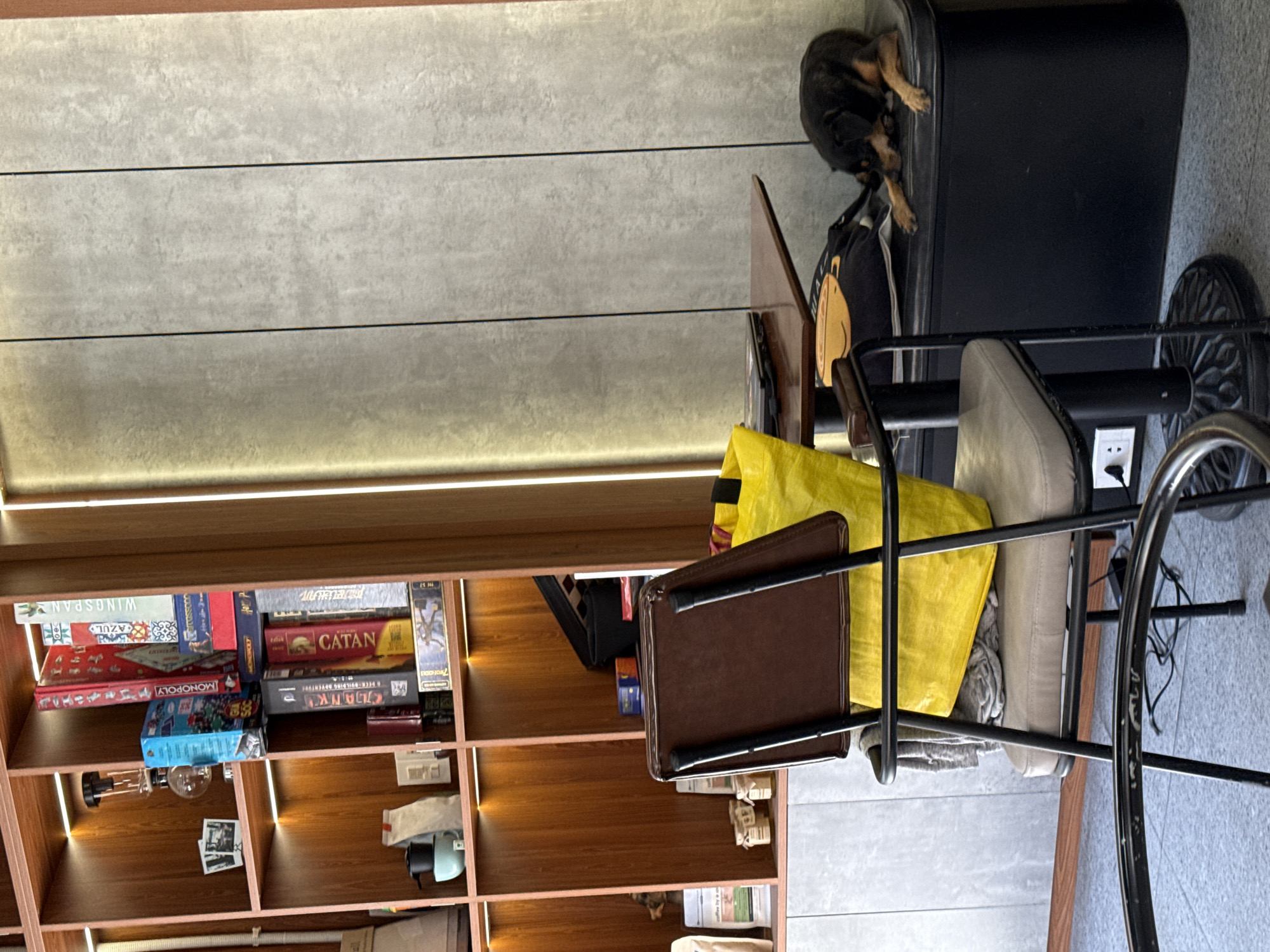The image shows an interior space in Đà Nẵng, Vietnam. A wooden bookshelf is filled with board games like "Wingspan," "Azul," "Monopoly," "Catan," and "Clank." A black metal frame chair with a brown seat and a bright yellow bag sits in the foreground. Atop a black cabinet next to the chair, a small dog is lying down. The wall behind the bookshelf is composed of gray, horizontal panels with recessed lighting. The floor is tiled with light gray squares.
