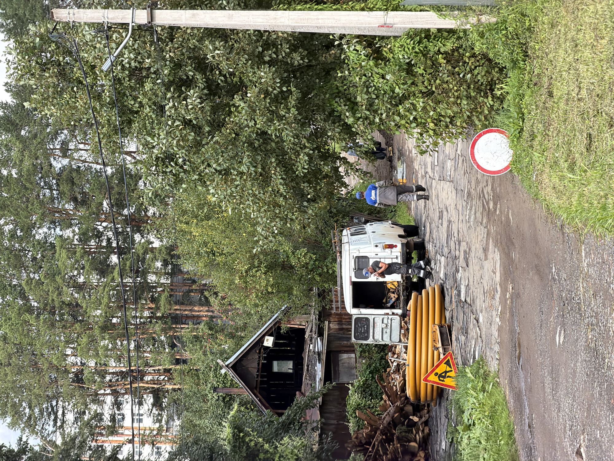In Chaikovsky, Russia, a rural dirt road, paved with cobblestones in some sections and marred by potholes and puddles, winds through a wooded area. On the left, a dilapidated wooden structure, possibly an abandoned house with a damaged roof, stands amidst a pile of cut logs. In front of the structure, a white van with its rear doors open is parked. Two men in work uniforms are present; one stands near the van's open doors, holding a mobile phone, while the other walks away from the camera, wearing a blue cap and a gray work suit with a "Gazprom" logo. Yellow, coiled pipes are stacked near the van, alongside a yellow triangular road sign with a depiction of a person digging. Further down the road, a circular traffic sign with a red border and a white center is visible. Lush green trees and foliage surround the scene, with tall pine trees dominating the background and a concrete utility pole to the right.
