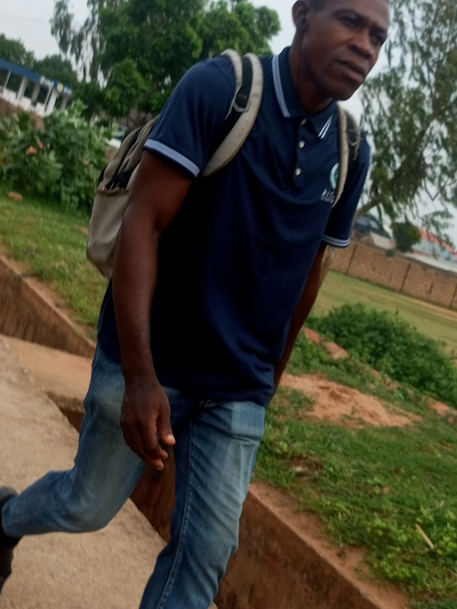 In Garko, Nigeria, a person is walking on a concrete staircase outdoors. The individual, wearing a navy blue polo shirt, blue jeans, and a light-colored backpack, is seen from the chest down, with their face partially visible in the upper right portion of the frame. They are looking slightly to the right. The background consists of green grass, dirt, and trees, with a building visible in the distance. The lighting suggests an overcast day.