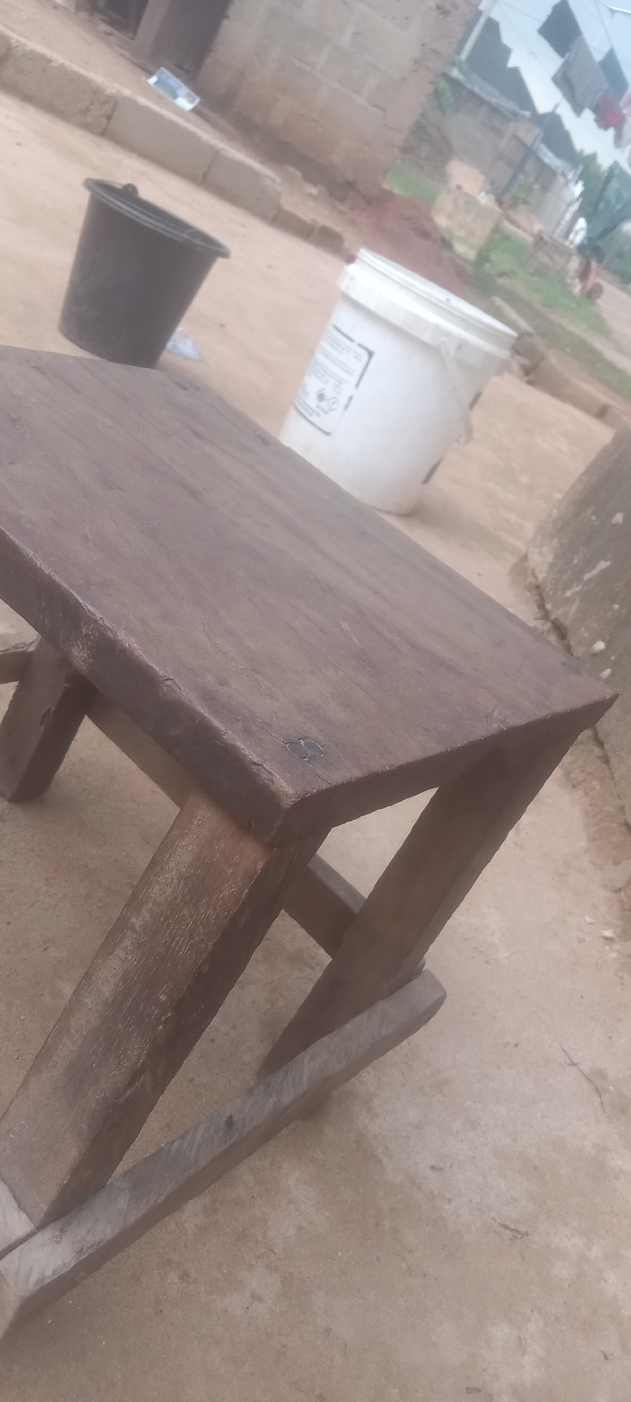 In a daytime outdoor setting in Bende, Nigeria, a handmade, dark wood, four-legged stool sits on a rough, tan concrete surface. Behind the stool are a black plastic bucket, a white plastic bucket, and an unidentified concrete structure. In the far background, a building under construction and clothes hanging on a line are visible. No people are near the stool.